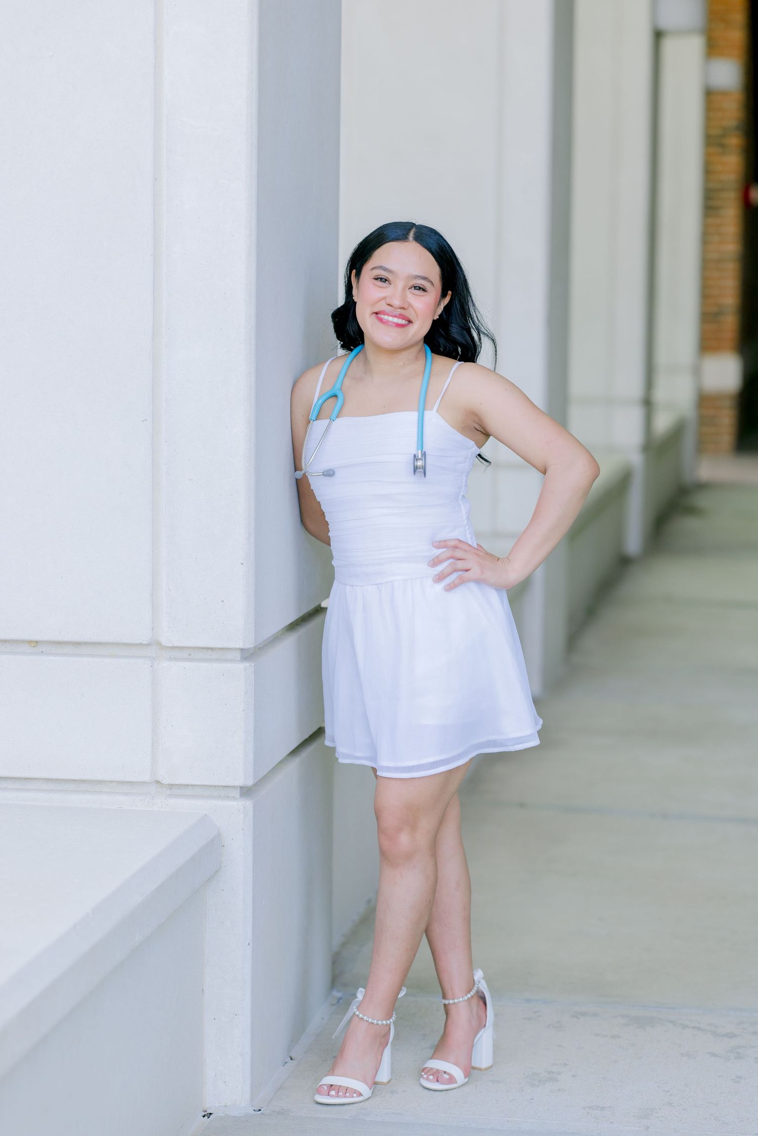 Young woman in white dress and heels with stethoscope poses confidently against a column outdoors.
