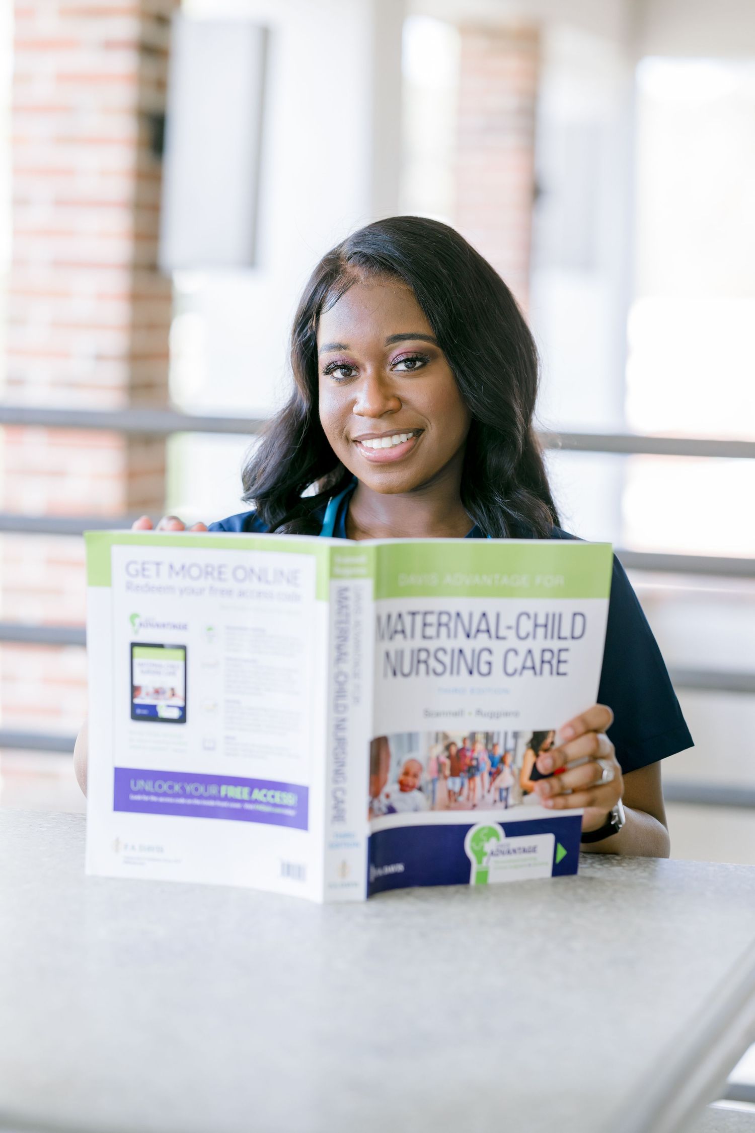 A nursing student studies behind an open textbook about maternal-child nursing care in a bright classroom setting.