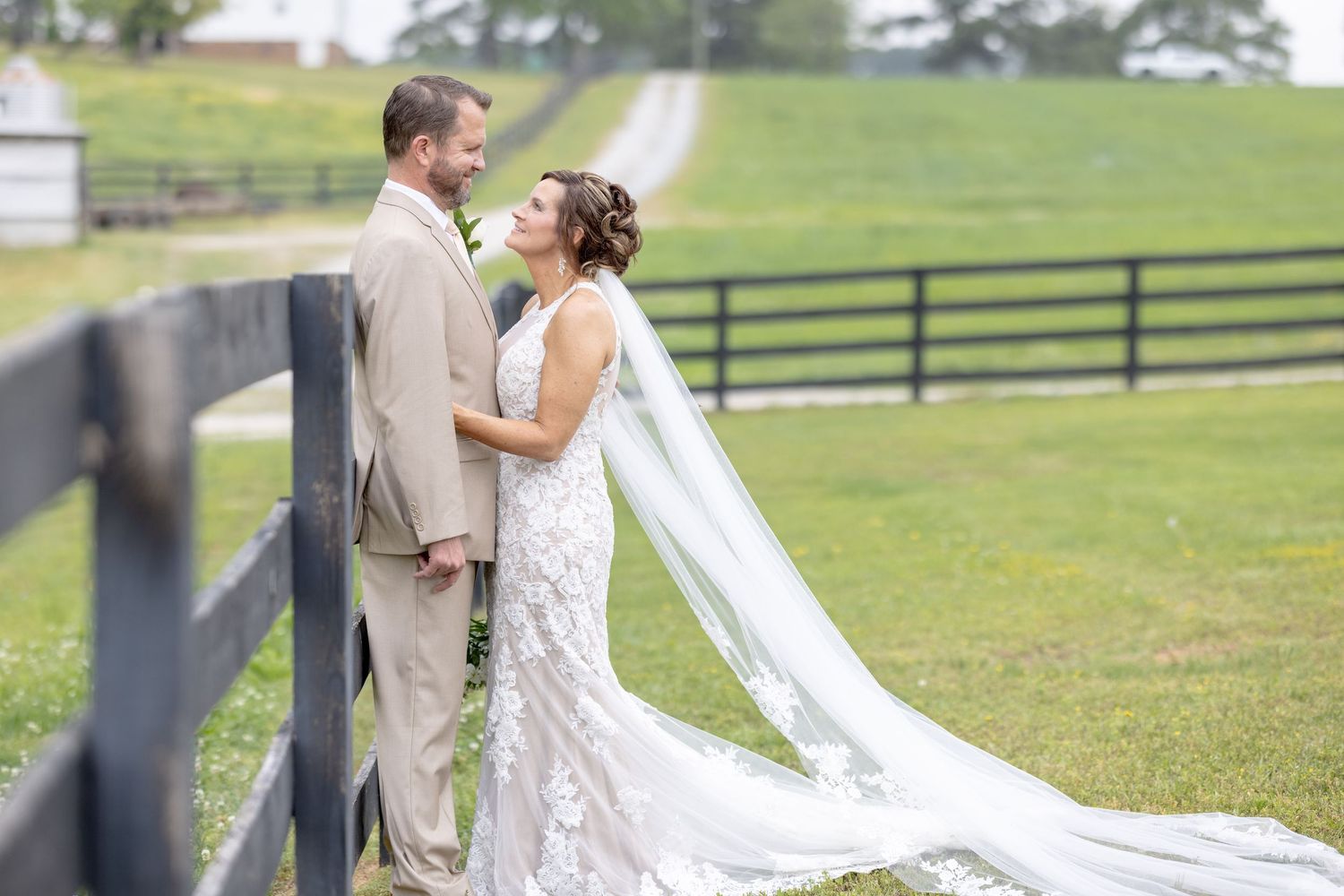 Bride and groom share a romantic moment by a wooden fence at a lush green farm wedding venue.