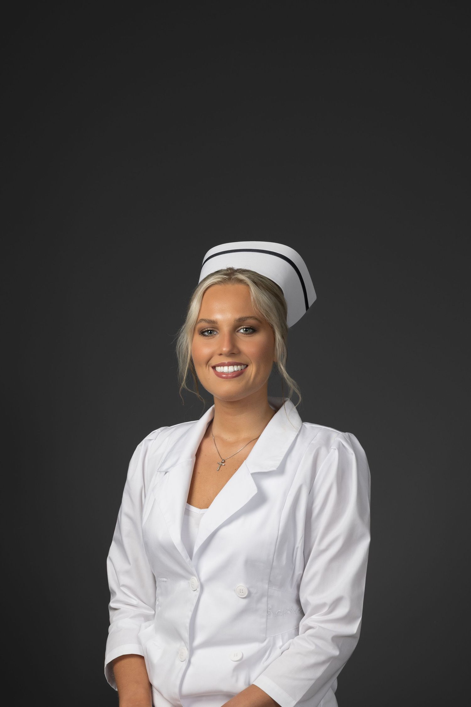 Smiling blonde woman in white nurse uniform and cap against dark gray background.