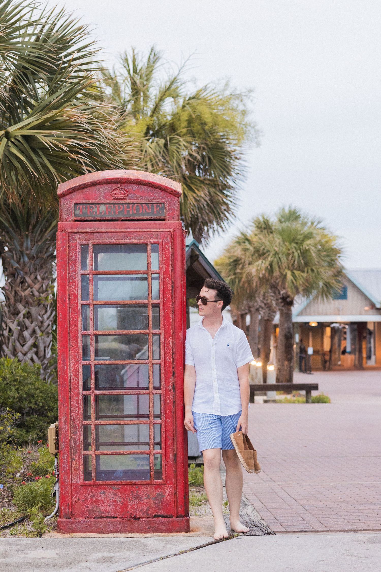 A vintage red British phone booth stands among palm trees on a coastal walkway.