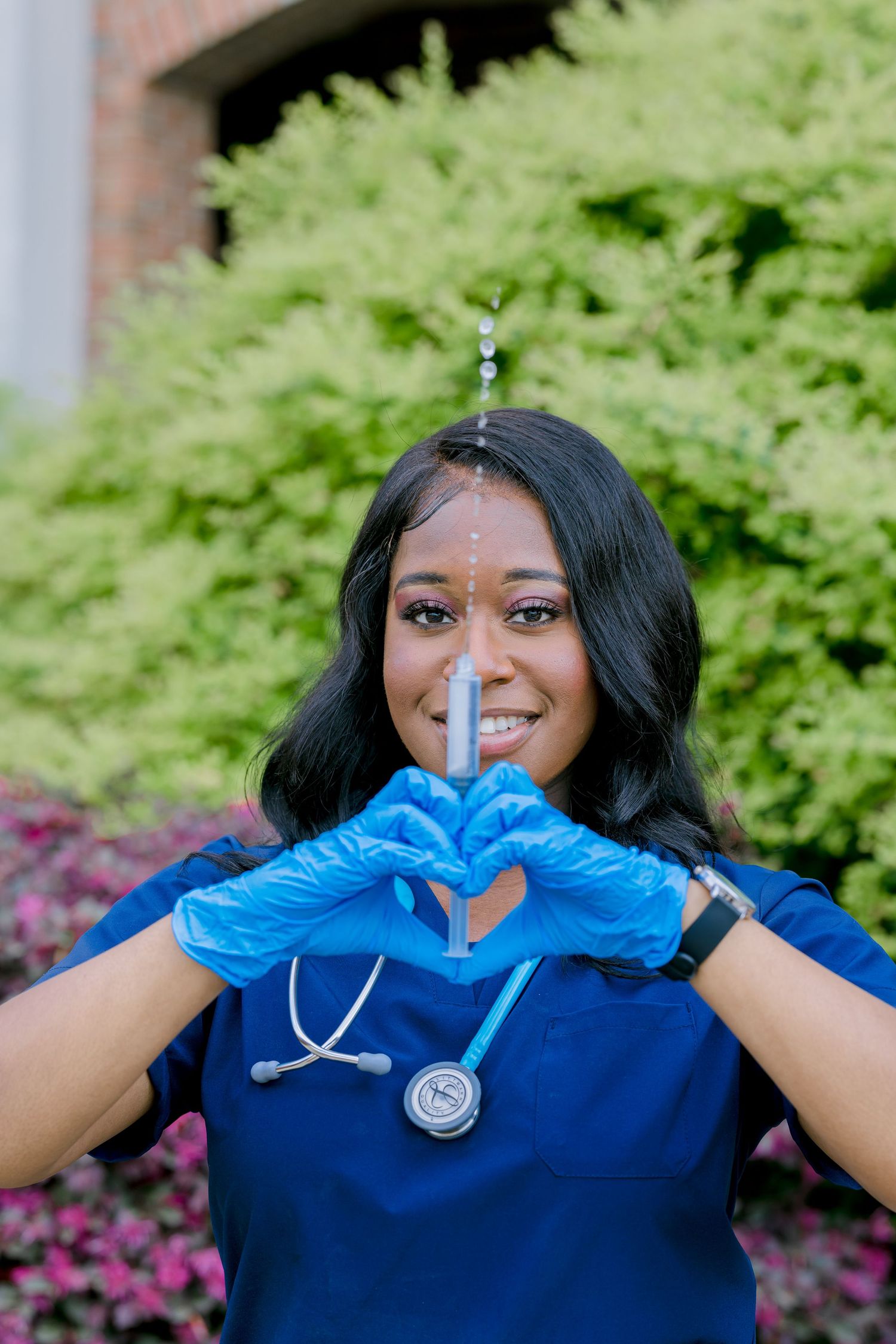 Healthcare worker in blue scrubs and gloves makes heart shape with hands while wearing stethoscope outdoors.