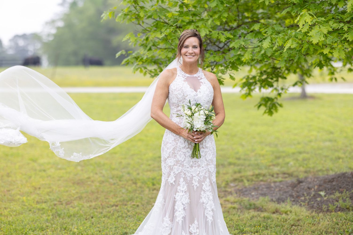 Bride in lace mermaid gown holding white bouquet with veil flowing in wind, standing outdoors under green tree.
