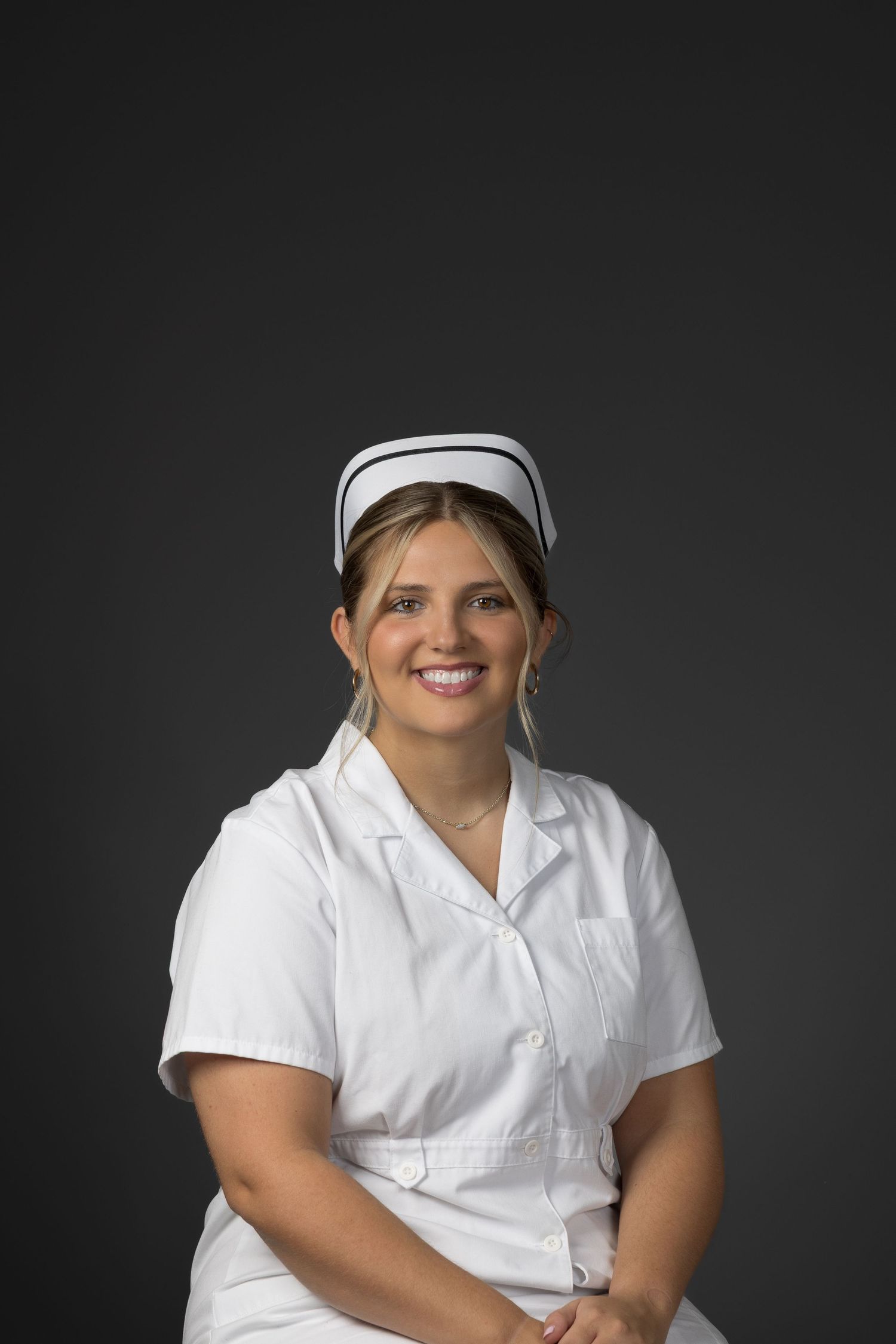 Smiling female nurse in white uniform and cap posed against dark gray background.