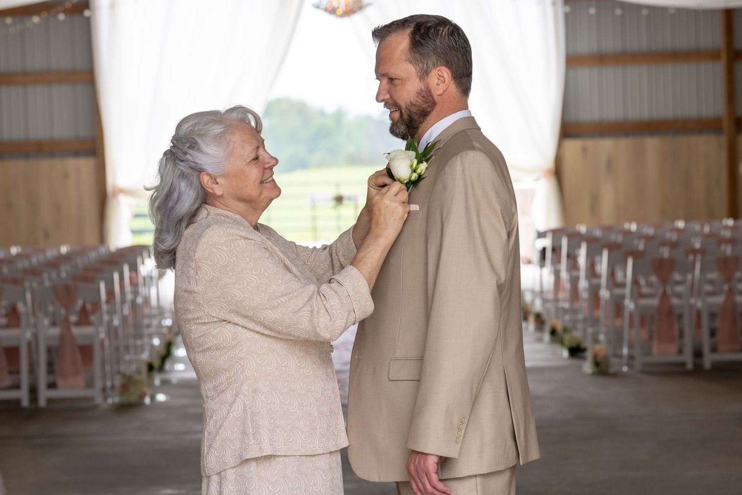 Elderly woman pinning a white boutonniere on a bearded groom in a tan suit inside a rustic barn wedding venue.