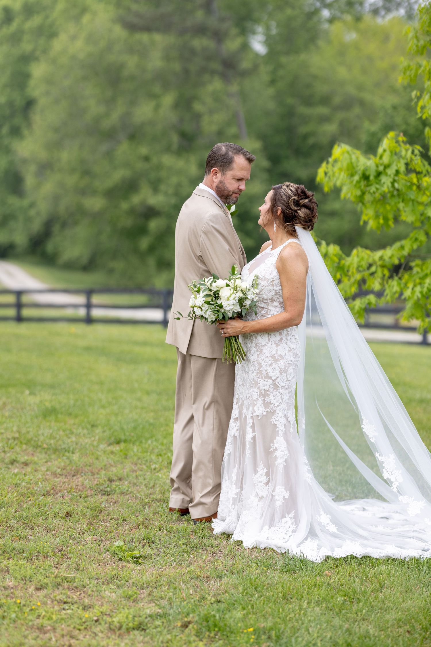 Bride and groom share a romantic moment outdoors, she in a lace gown with veil, he in a tan suit, surrounded by greenery.