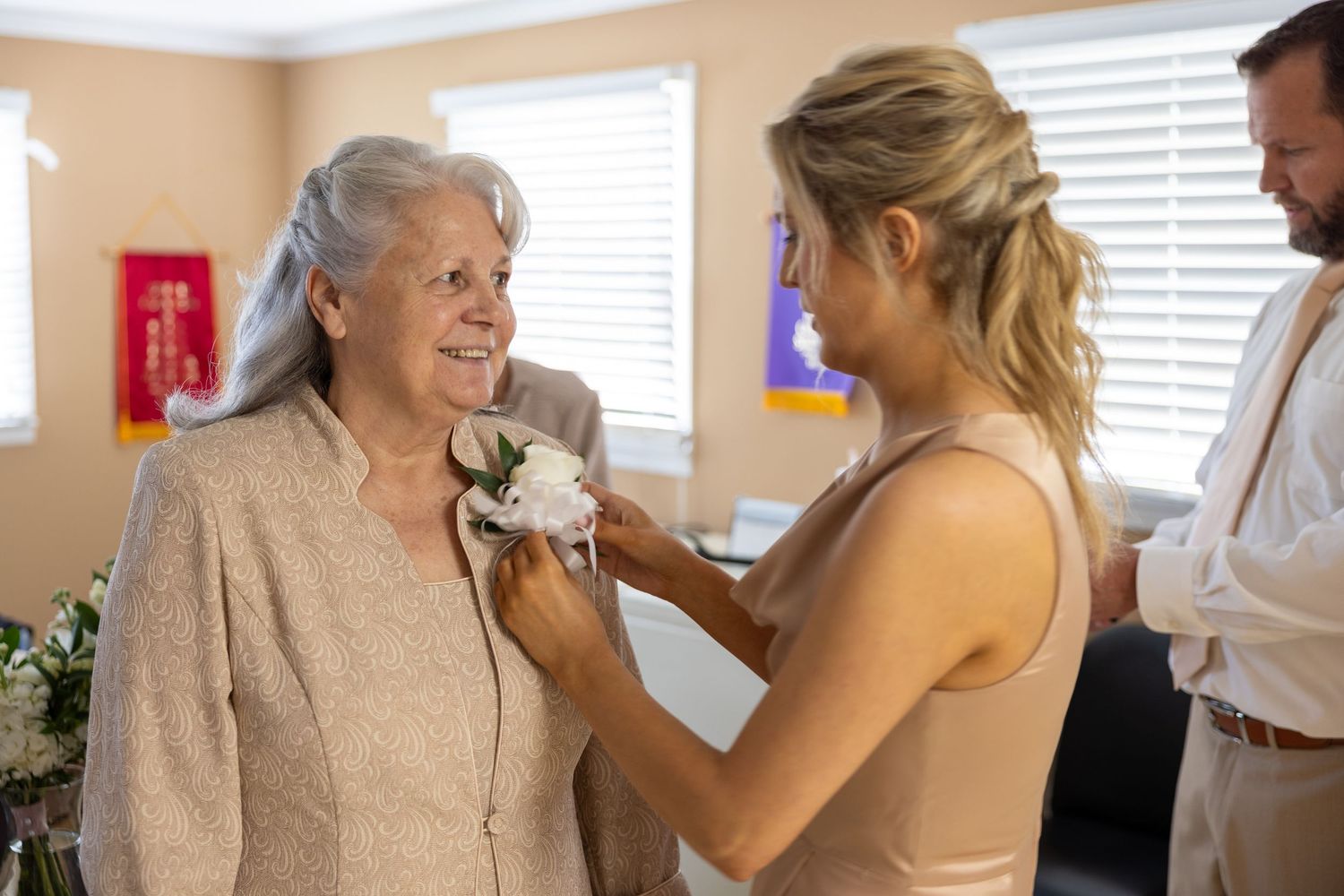 Young woman pinning a white corsage on an older woman's lapel at a wedding celebration indoors.