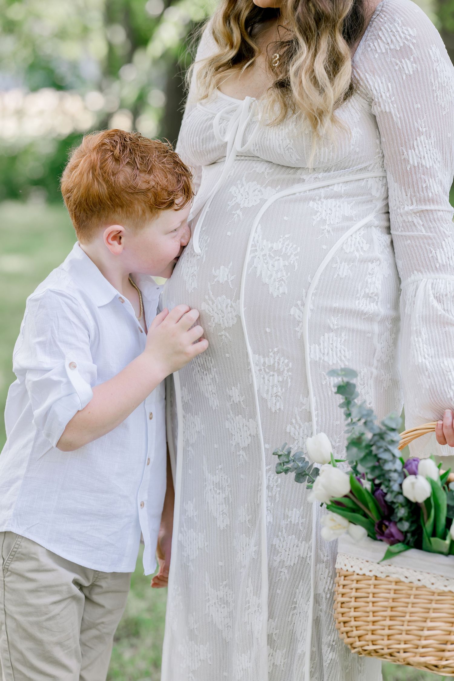Red-haired boy kissing pregnant mother's belly outdoors, mom in white lace dress holding flower basket.