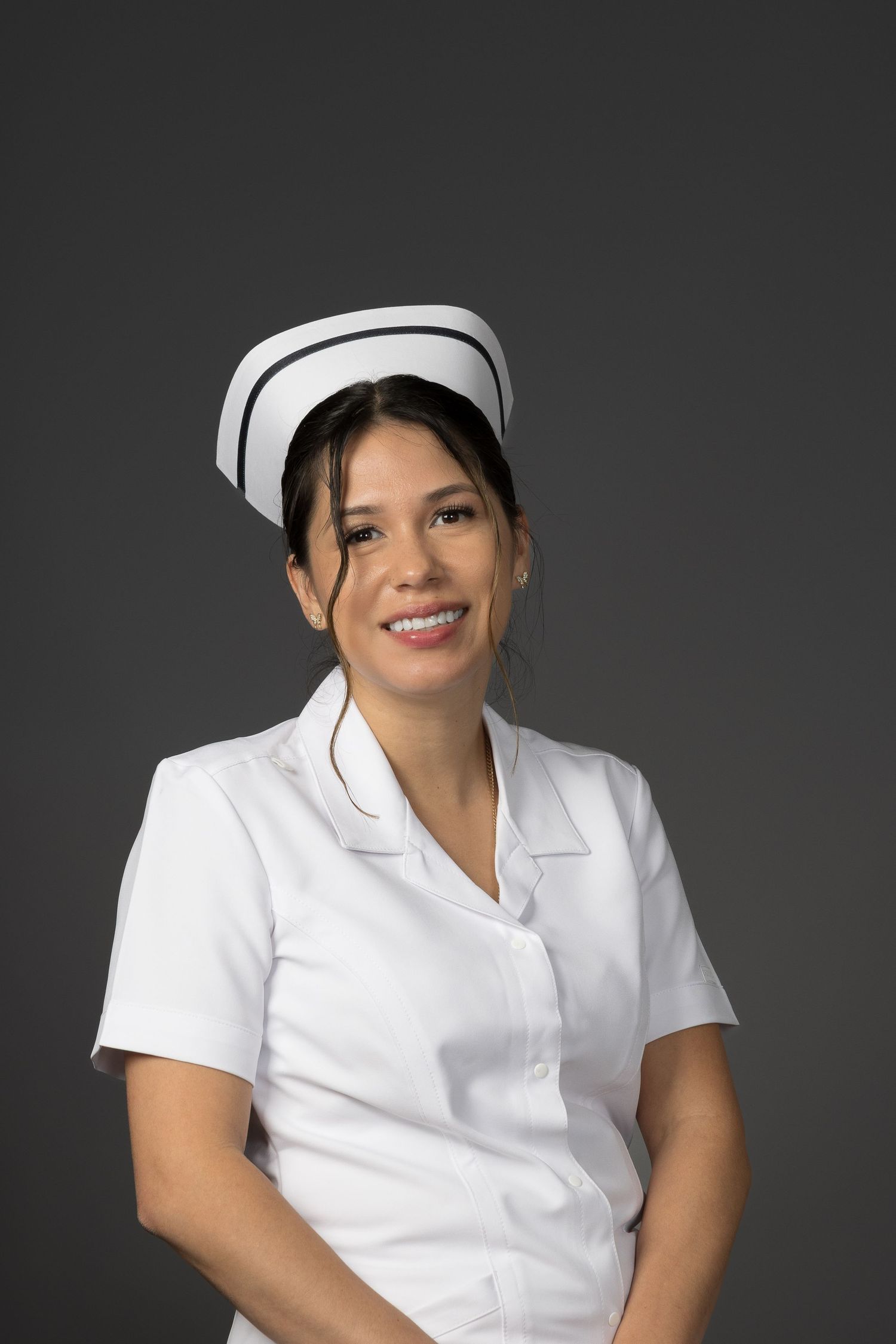 Professional female nurse in white uniform and cap smiling confidently against a gray background.