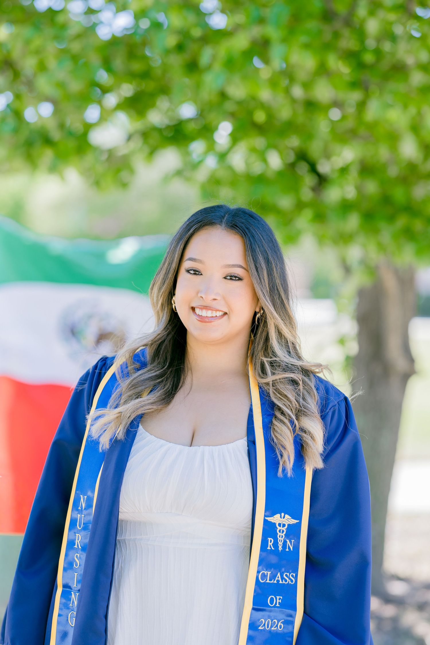 Smiling nursing graduate in blue cap and gown with RN Class of 2026 stole, posing outdoors near Mexican flag.