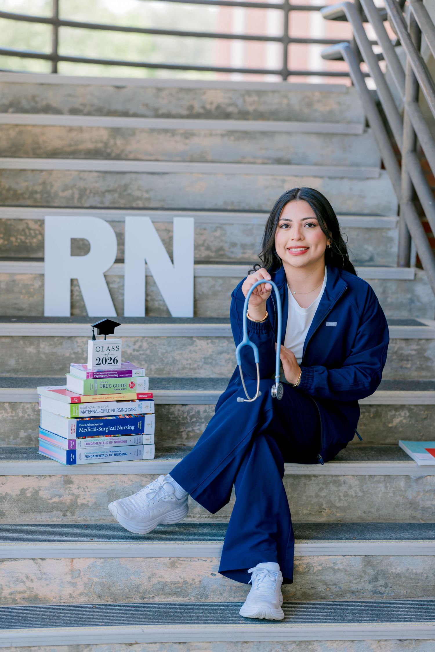 Nursing student in navy scrubs poses on steps with RN letters, stethoscope, and Class of 2026 textbooks.
