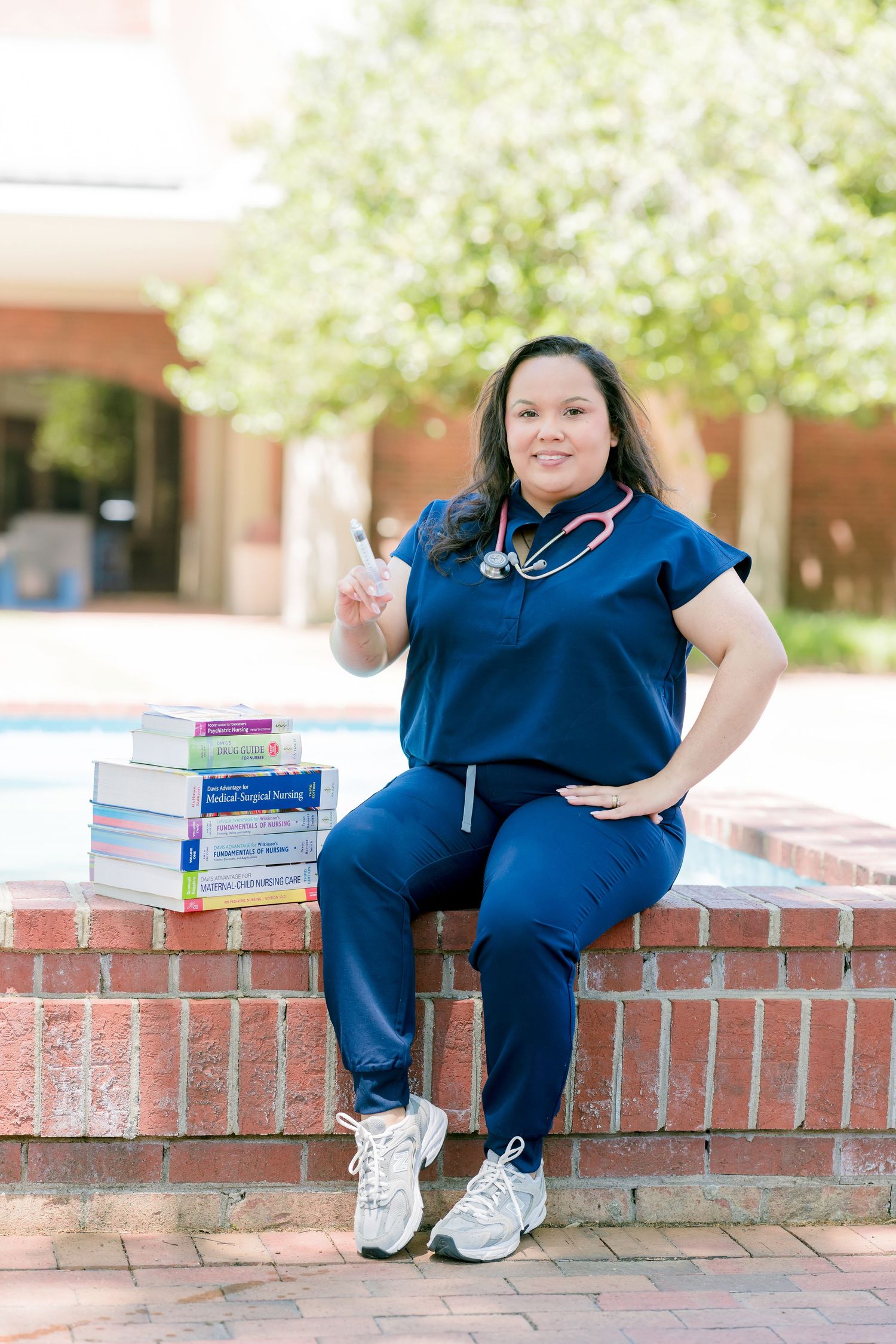 Nursing student in blue scrubs with stethoscope sitting on brick wall beside stacked medical textbooks, flashing peace sign.