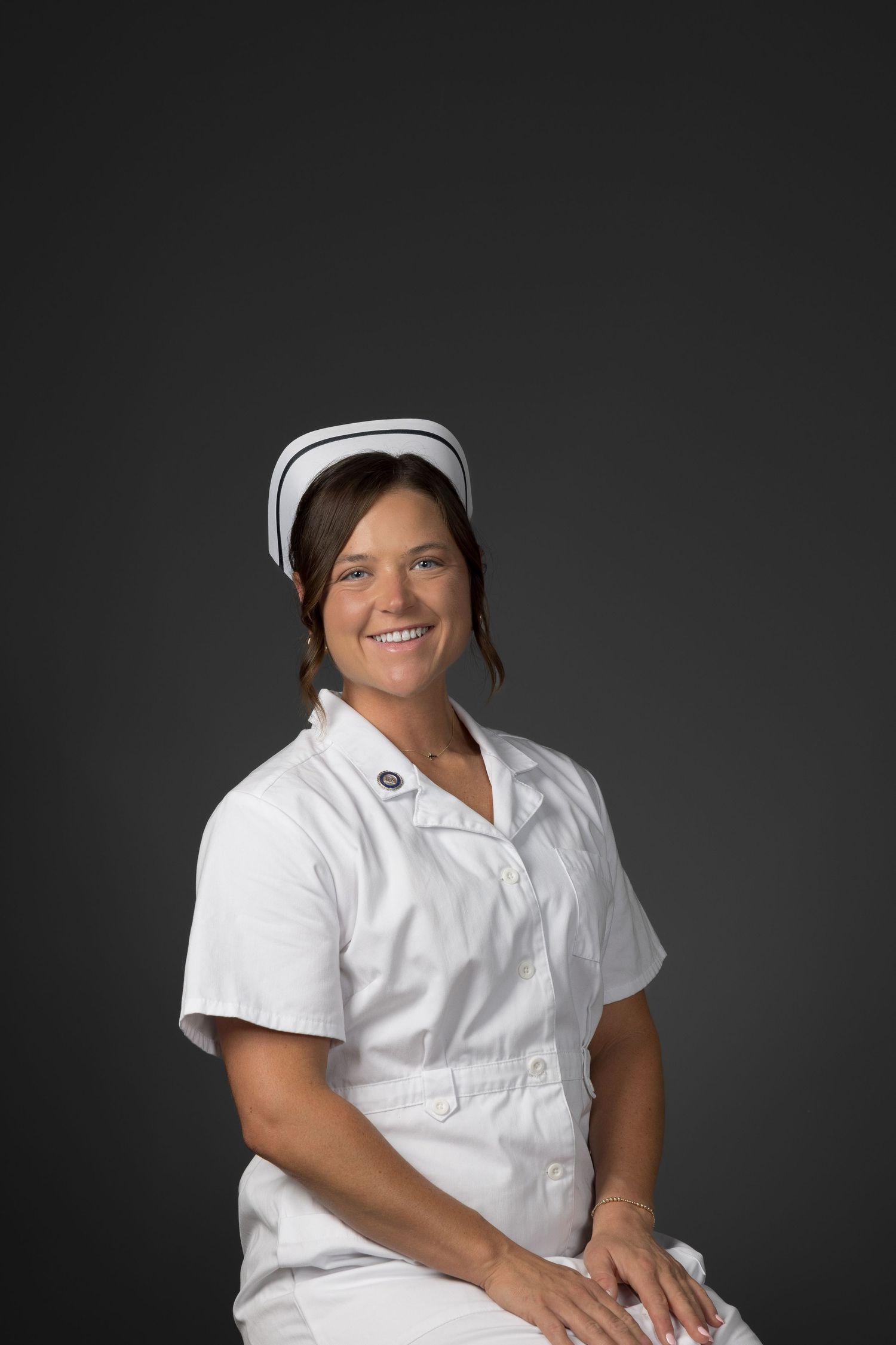 Smiling female nurse in white uniform and cap seated against dark gray background.