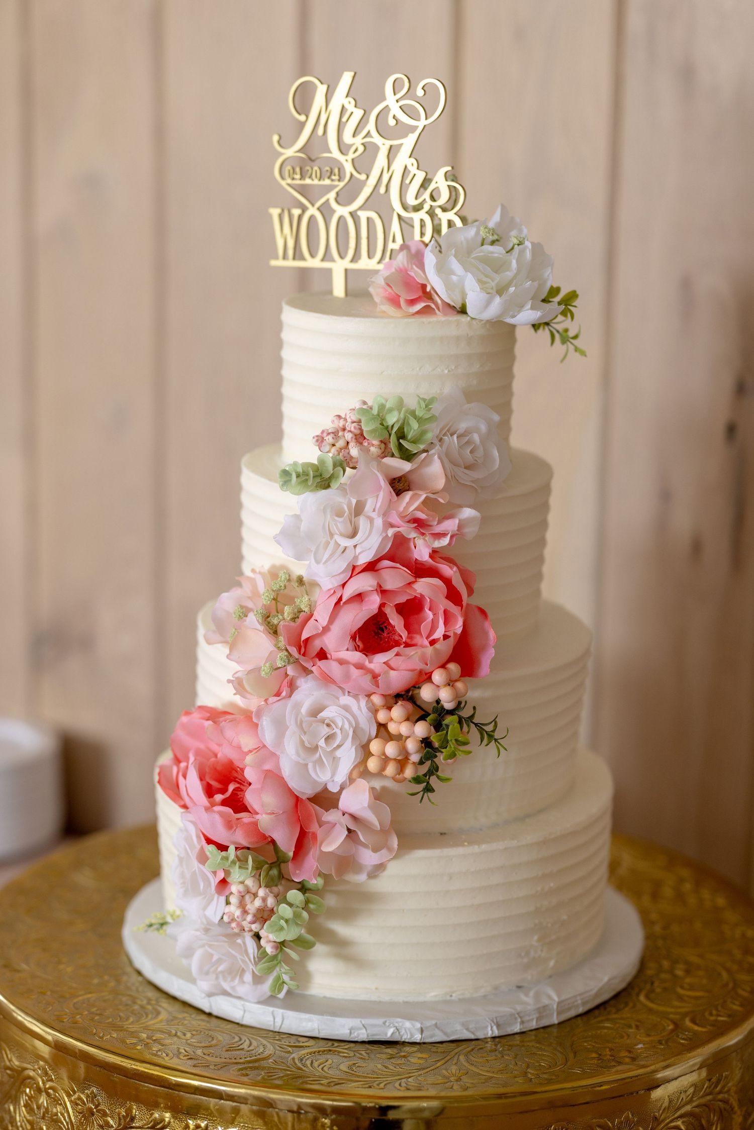 Three-tier white wedding cake with pink peonies, roses, and a gold Mr. & Mrs. Woodard cake topper.