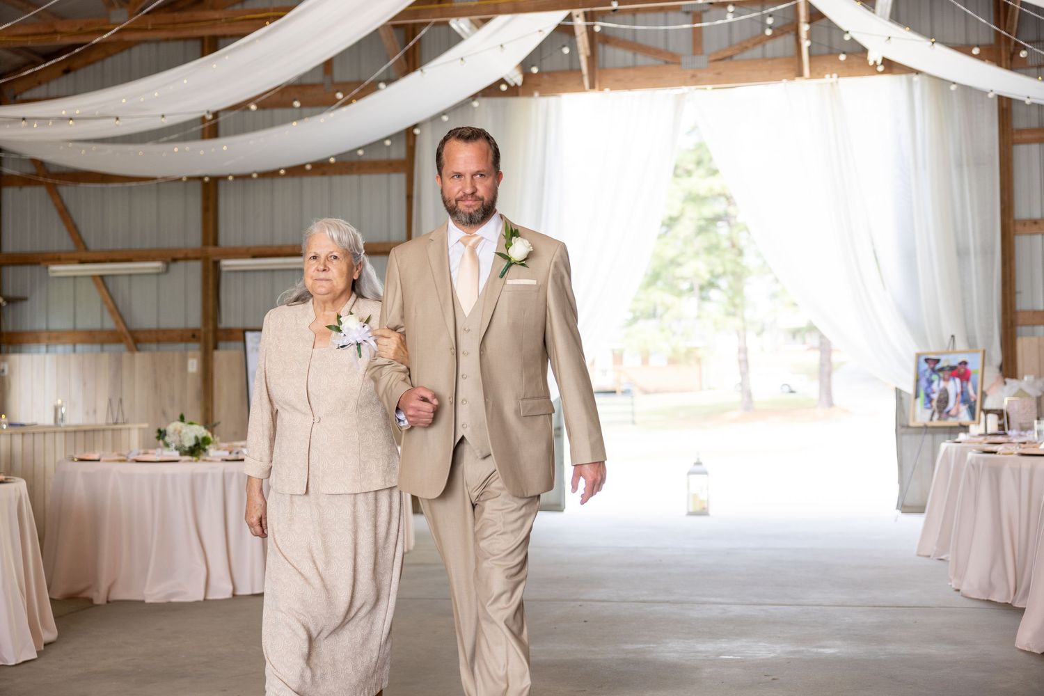 Groom in tan suit escorts elderly woman in beige pantsuit down barn wedding aisle with white drapes.