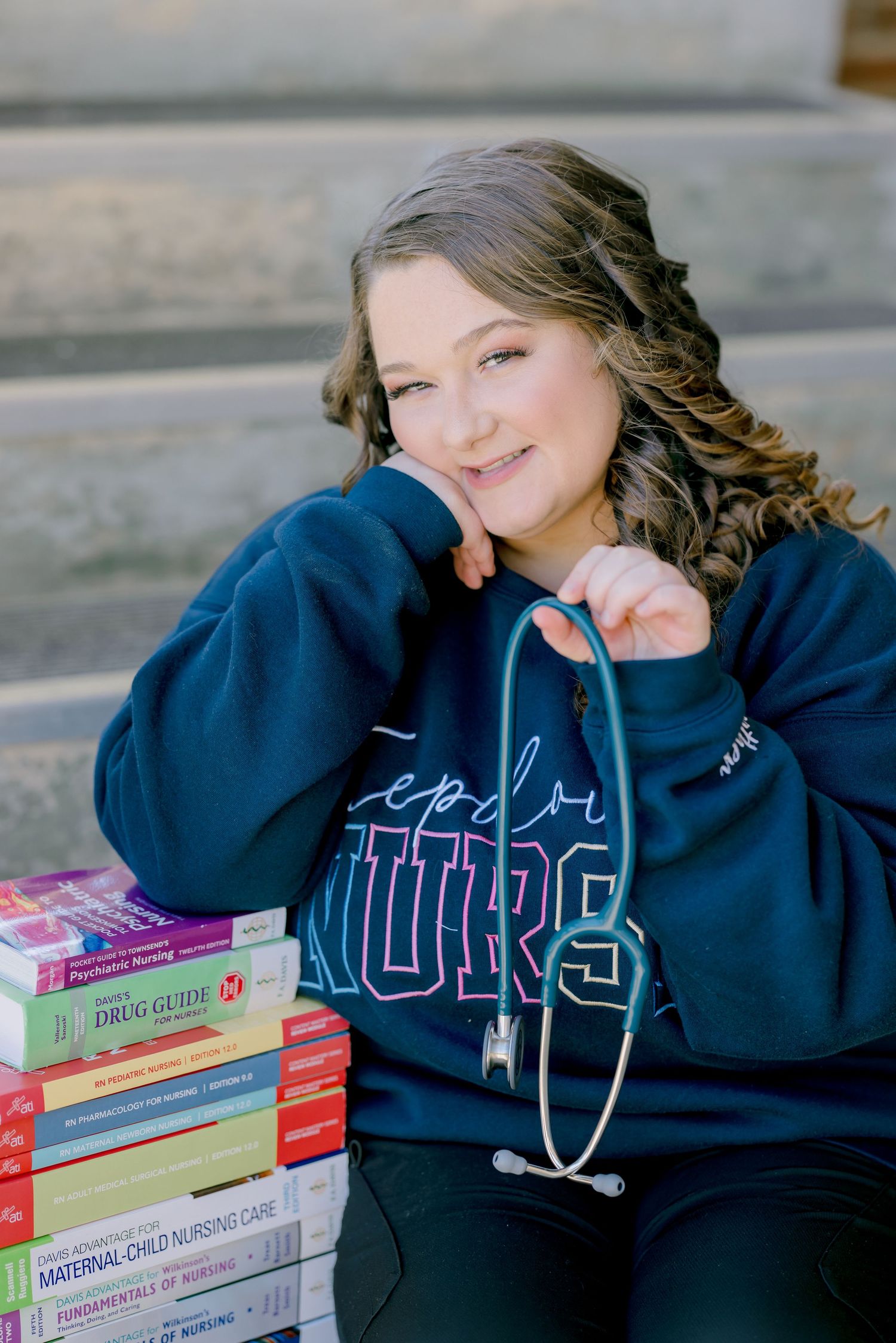 Smiling nursing student holding stethoscope beside stack of nursing textbooks wearing navy Future Nurse sweatshirt on steps.