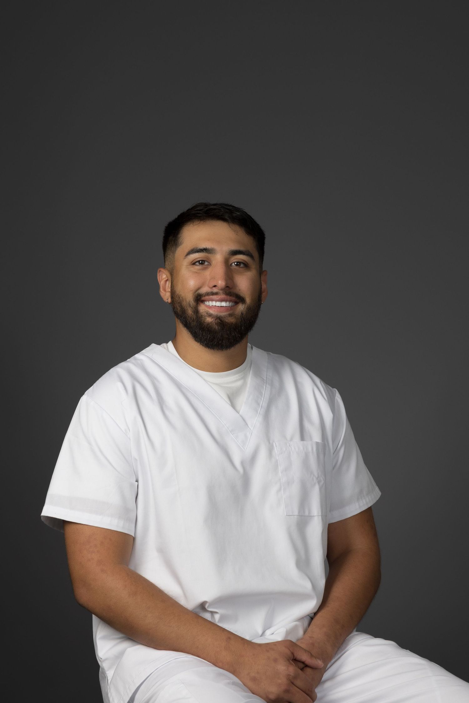 Bearded male healthcare worker in white scrubs smiling warmly against gray background.