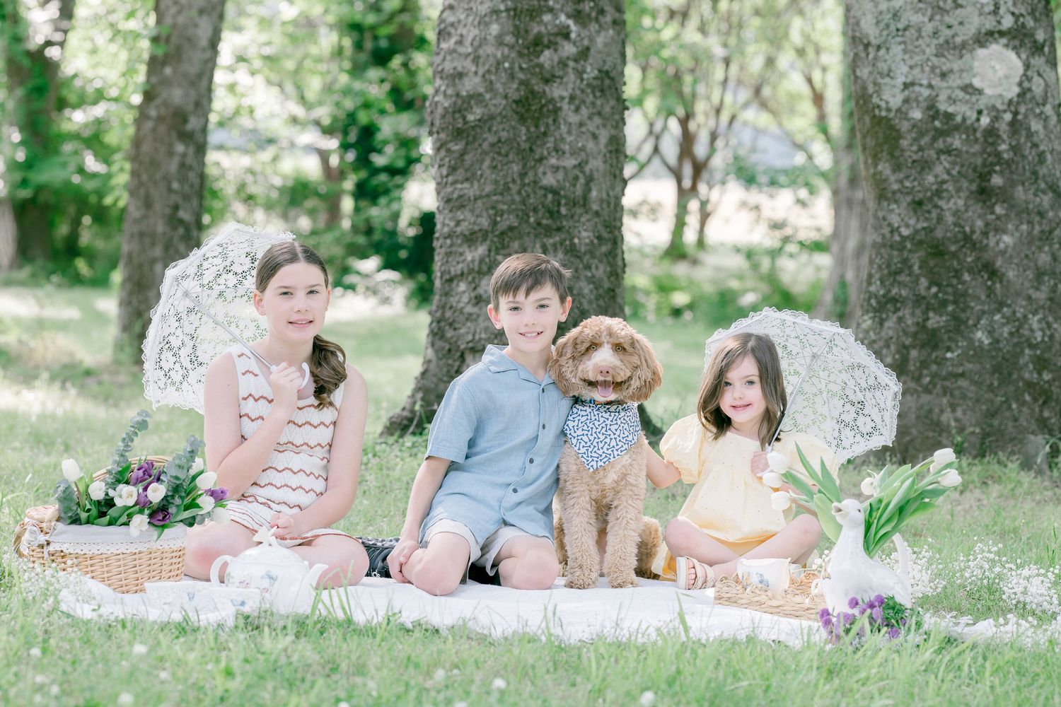 Three children and a golden doodle dog enjoy a sunny outdoor picnic on a white blanket surrounded by lush green trees.