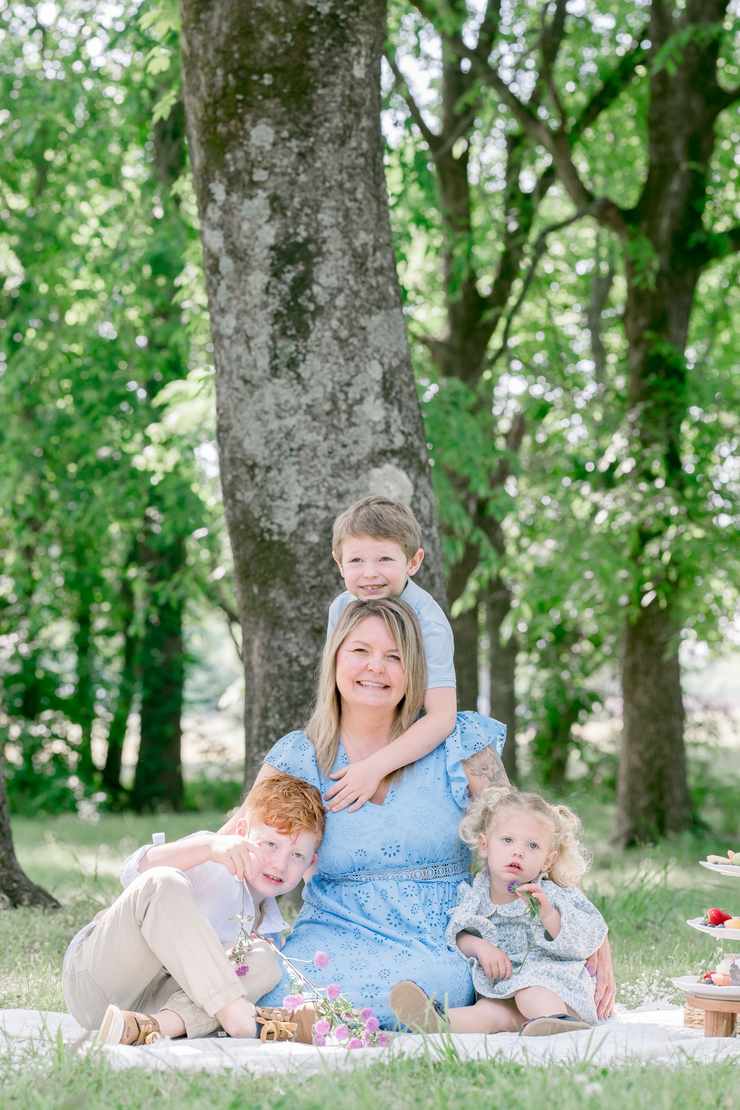 A smiling blonde mother in a blue dress sits on a blanket outdoors with her three young children under large green trees.