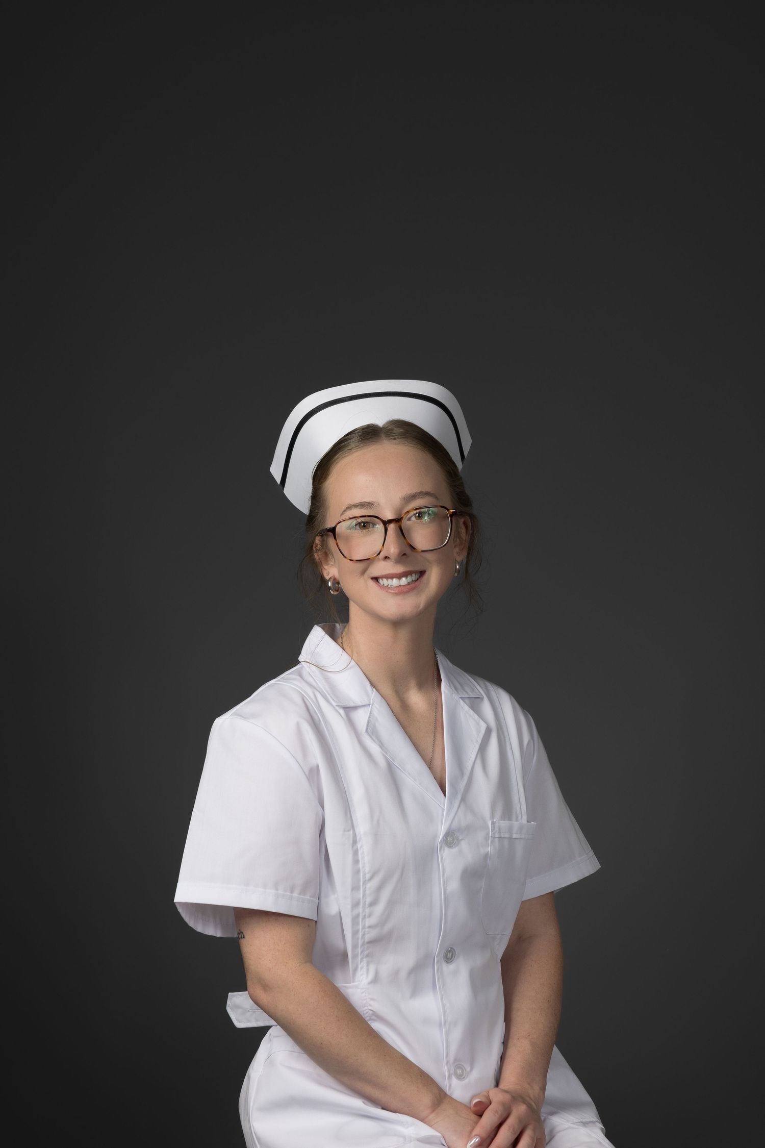 Young female nurse wearing glasses and white uniform with cap, smiling against gray background.