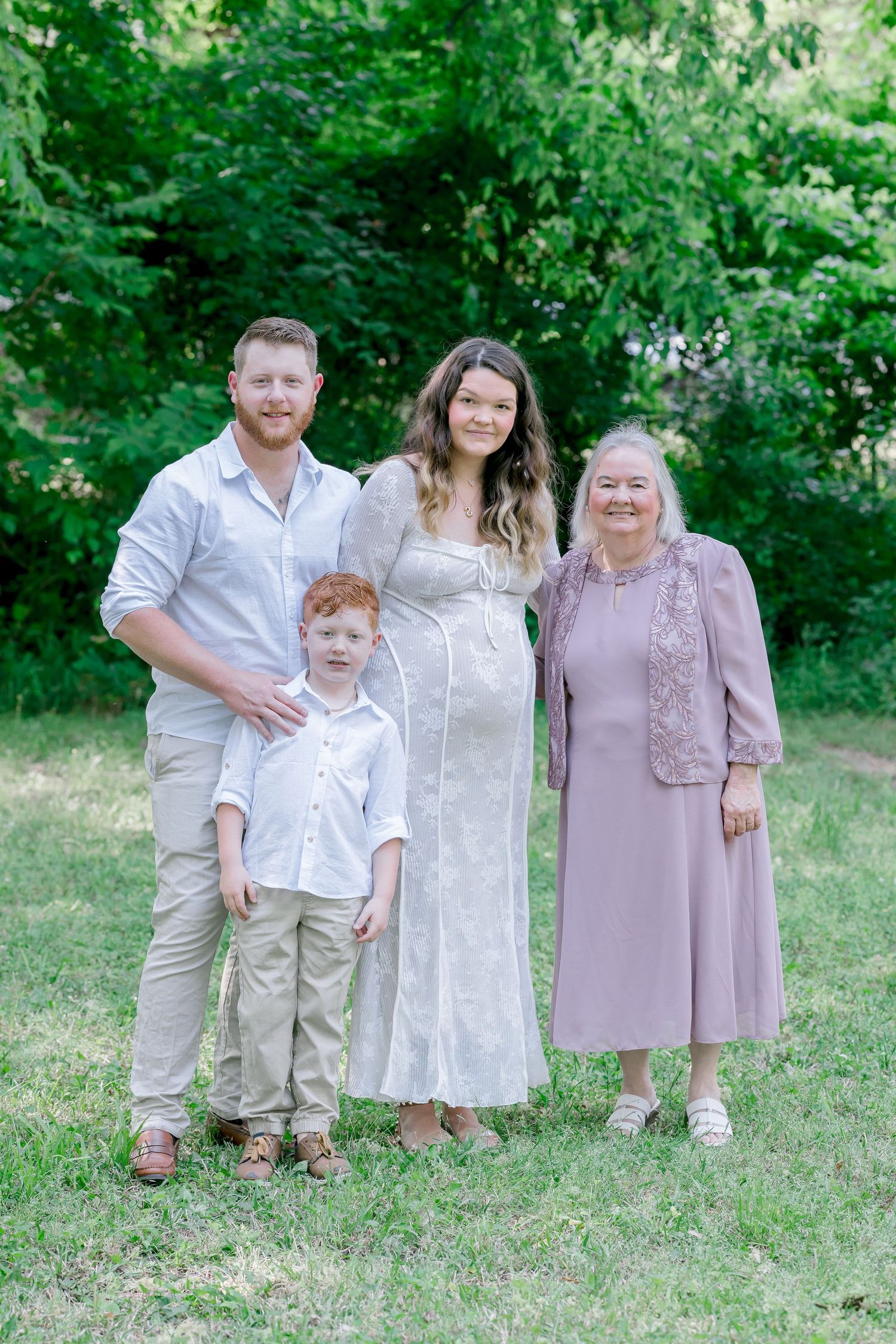 Family of four posing outdoors in coordinating outfits, including a pregnant woman in white dress, on green grass.