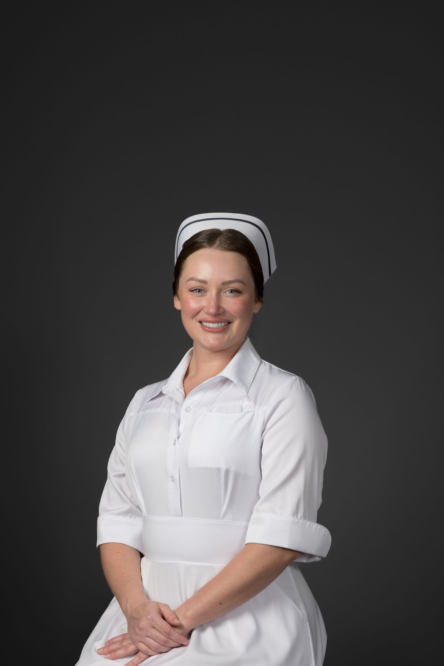 Cheerful brunette nurse in traditional white uniform and cap smiling against gray background.