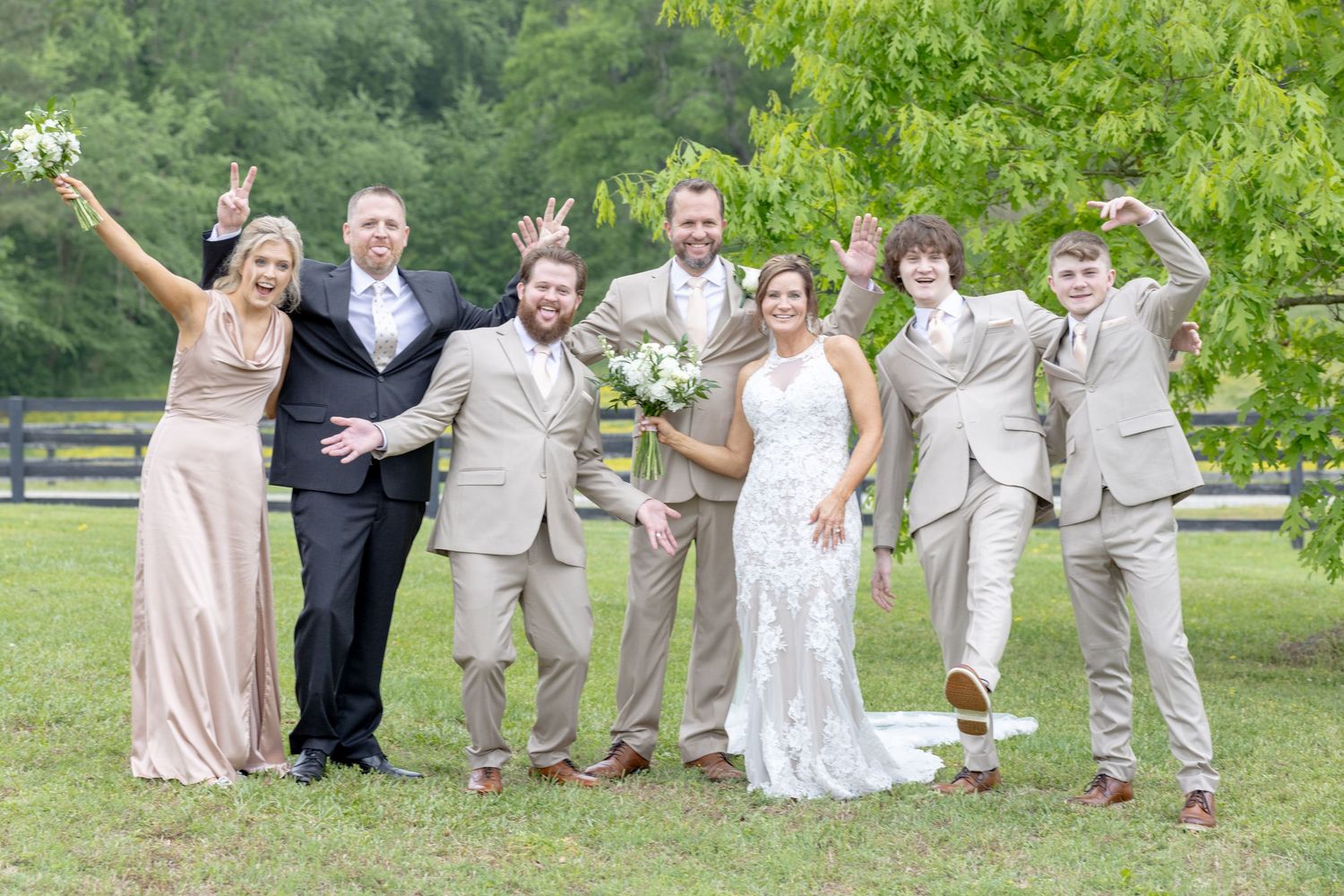 Wedding party posing playfully outdoors on green grass, bride in lace gown with bouquet, groomsmen in tan suits, bridesmaid in blush.