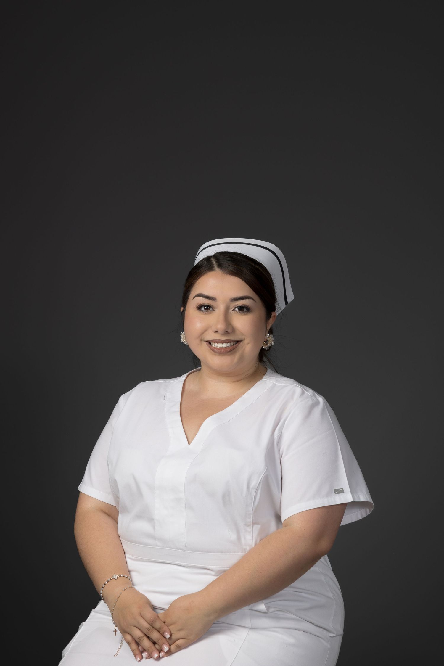 Cheerful female nurse in white scrubs and cap sitting with hands folded against dark background.