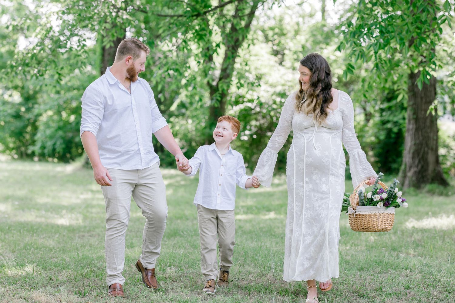 Pregnant woman in white lace dress holds basket of flowers while walking with husband and young red-haired son in green park.