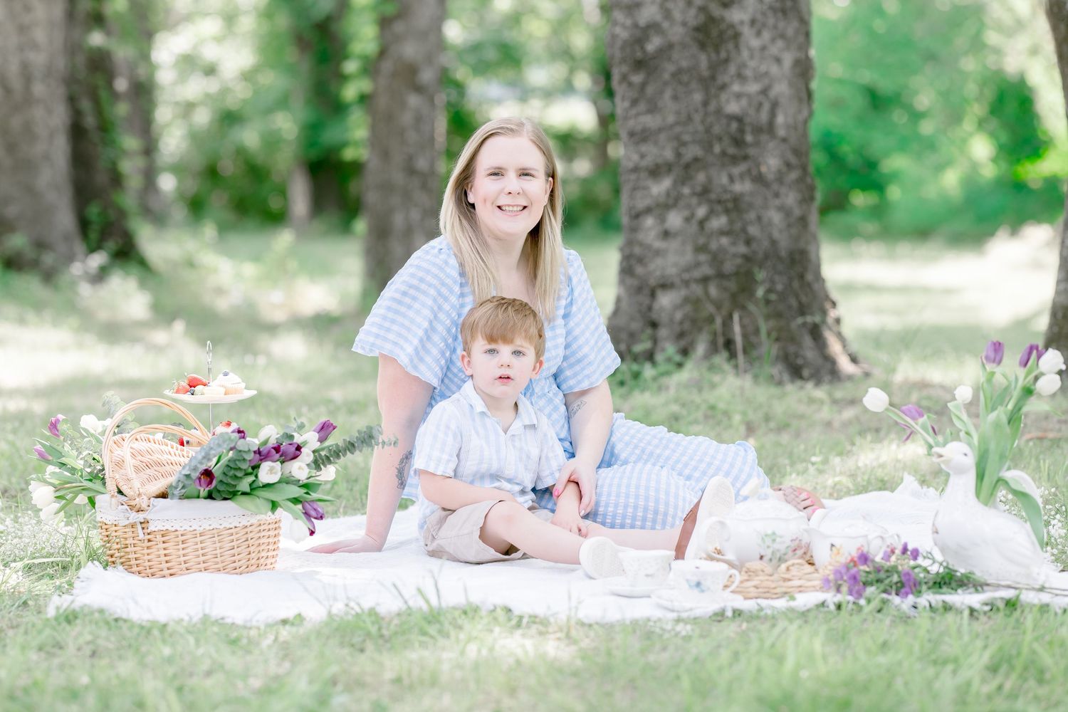 A mother and toddler son sit together on a white blanket for a picnic under trees in a lush green park.