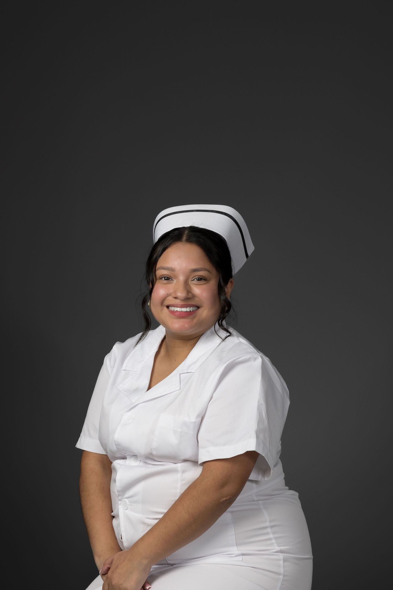 Smiling female nurse in white uniform and cap posing against gray background.