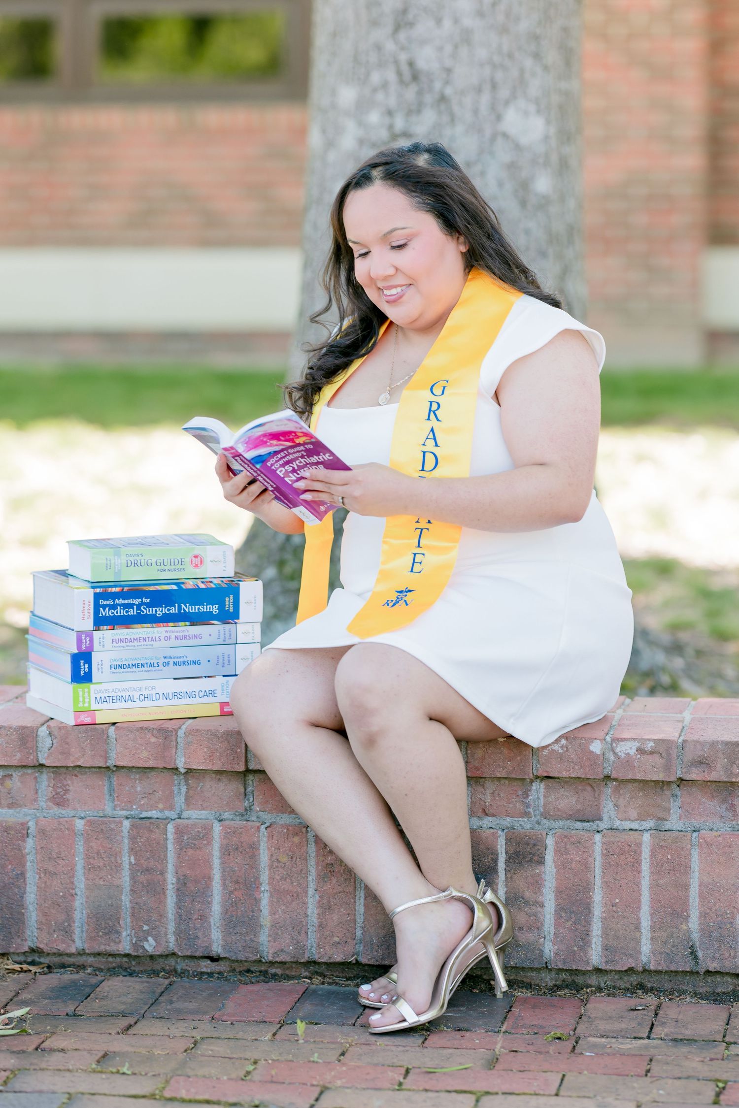 Nursing graduate in white dress and gold sash reads textbook beside stacked medical books on brick wall.