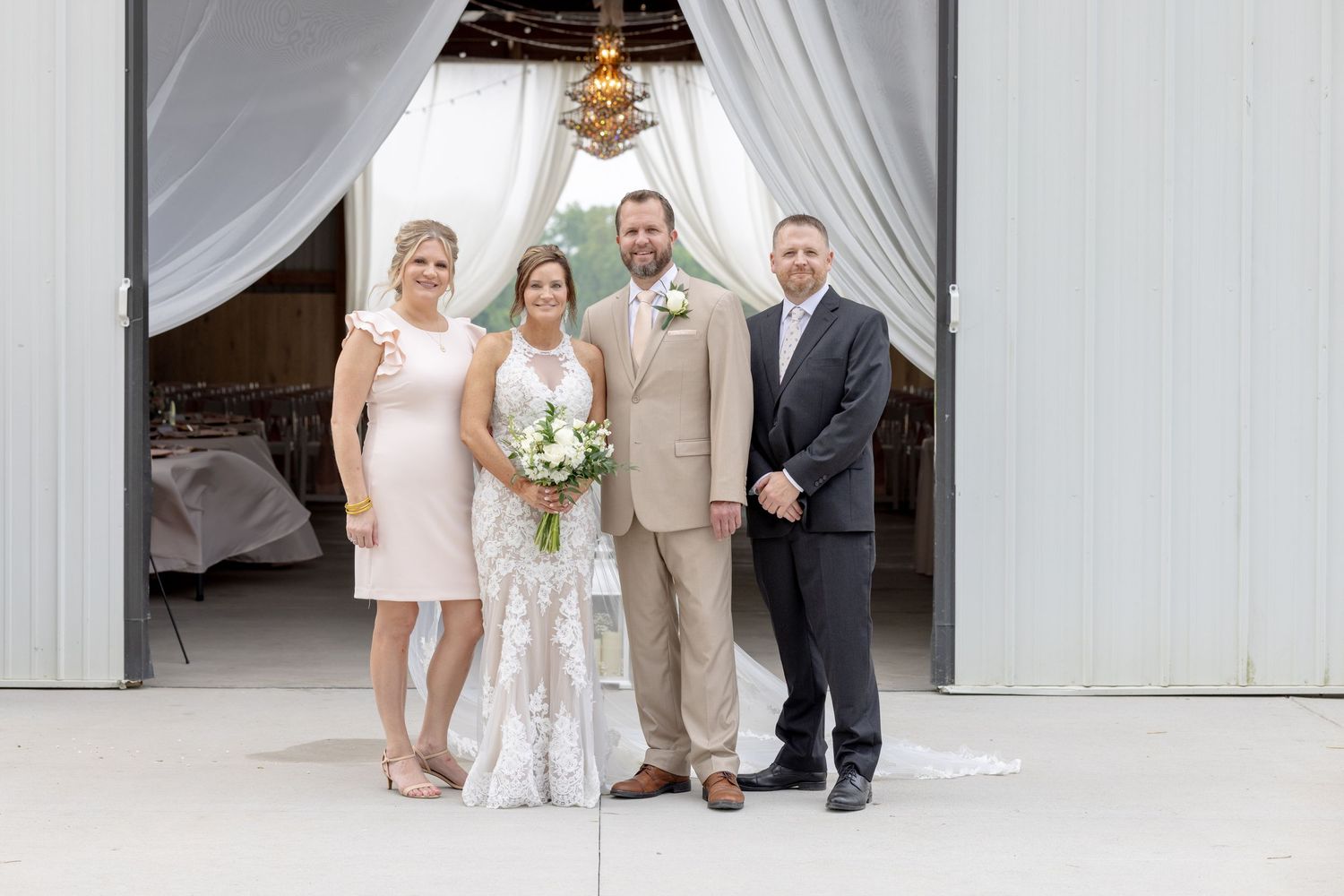 Bride and groom pose with two guests at a rustic barn wedding venue decorated with white draping and floral arch.