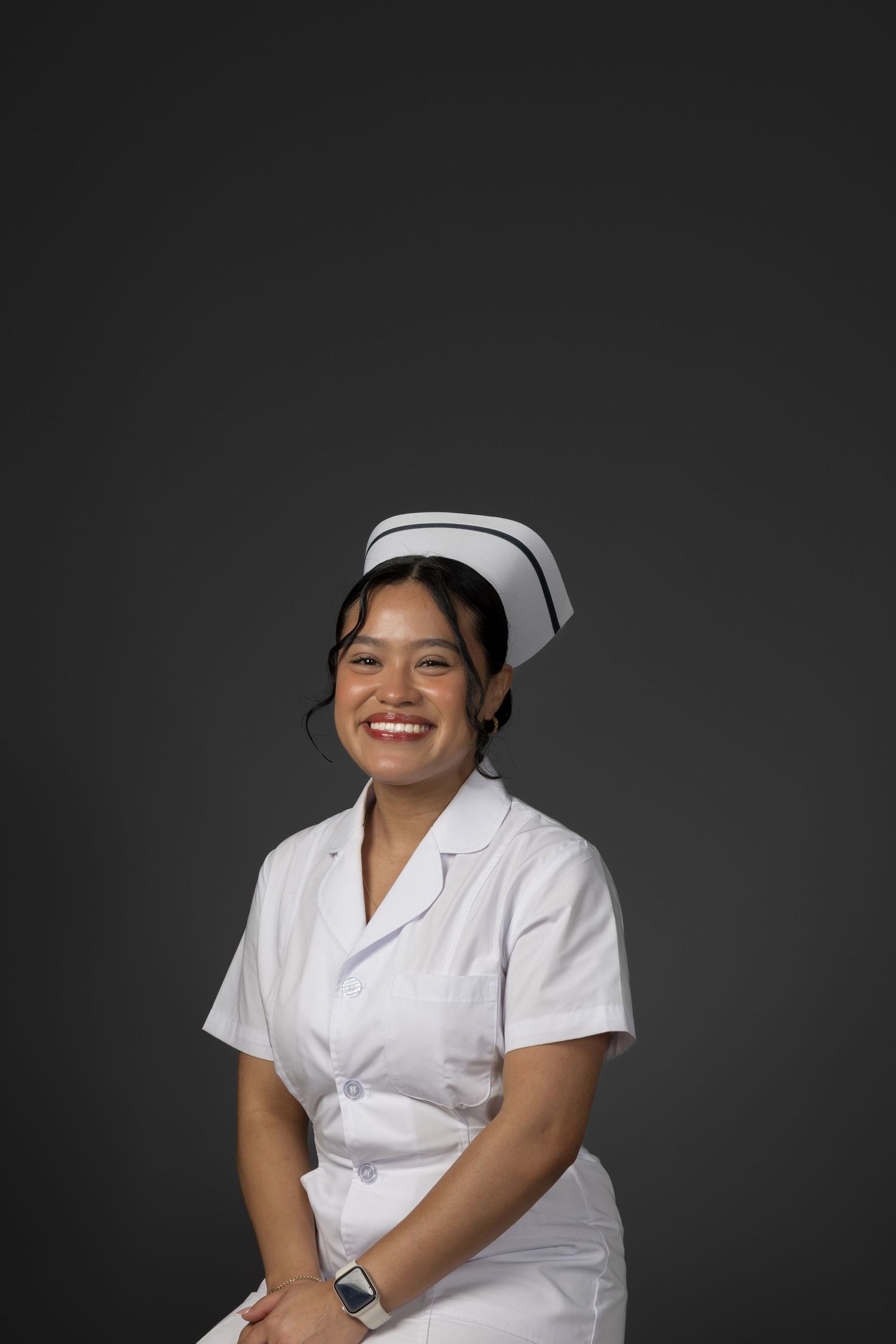 Happy young Asian female nurse in white uniform and cap laughing against gray background.