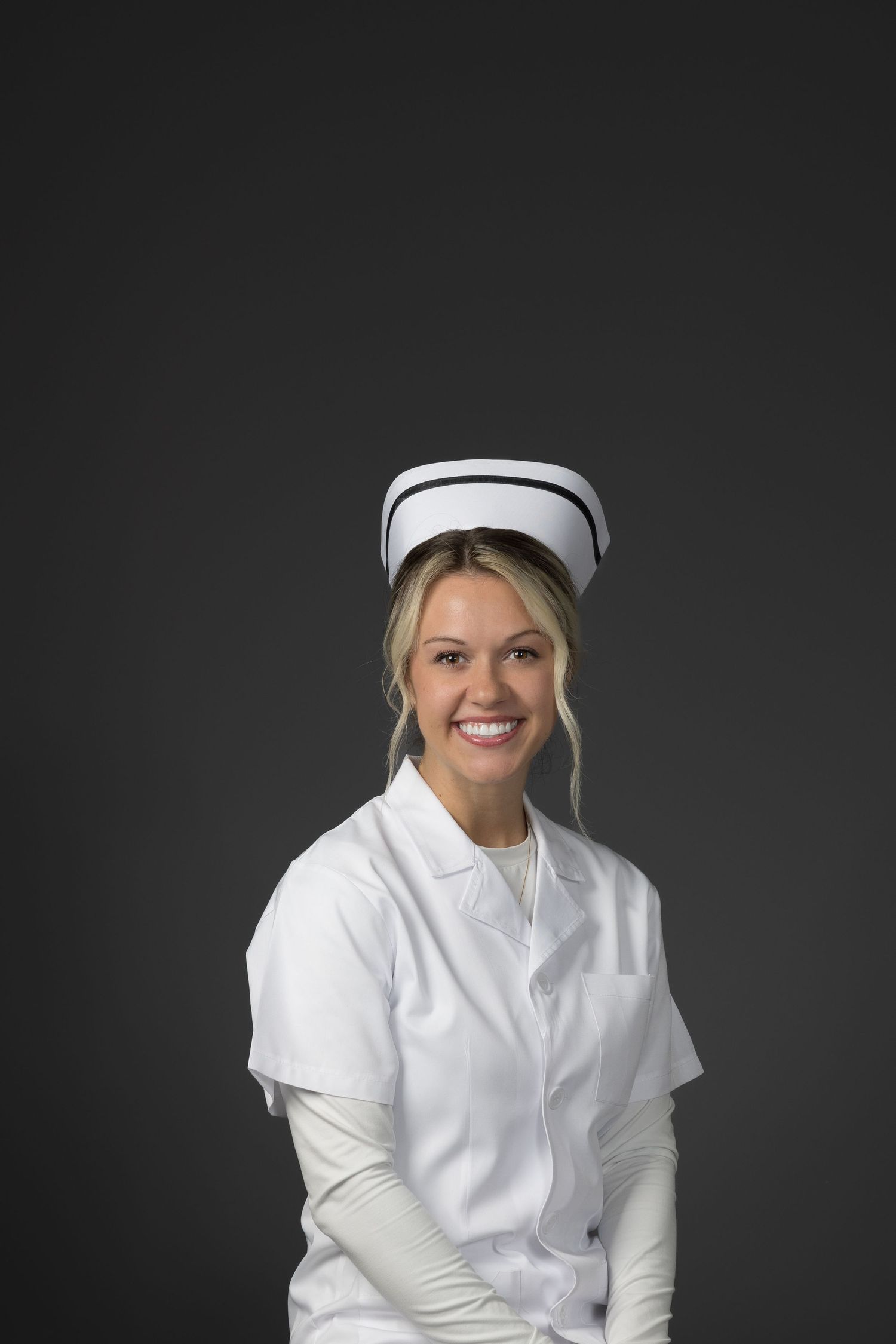 Smiling blonde female nurse in white uniform and traditional cap posing against a dark gray background.