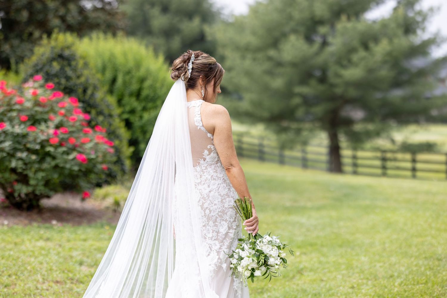 Bride in lace wedding dress and veil holding white bouquet on green lawn with red roses and fence.