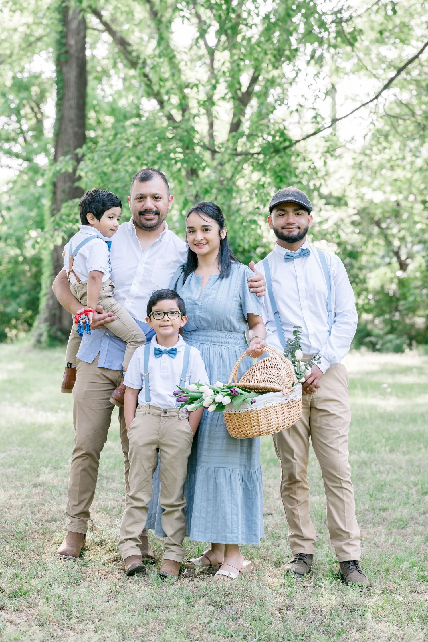 Family of five posing outdoors in coordinated blue and khaki outfits, holding a flower basket in a lush green park.