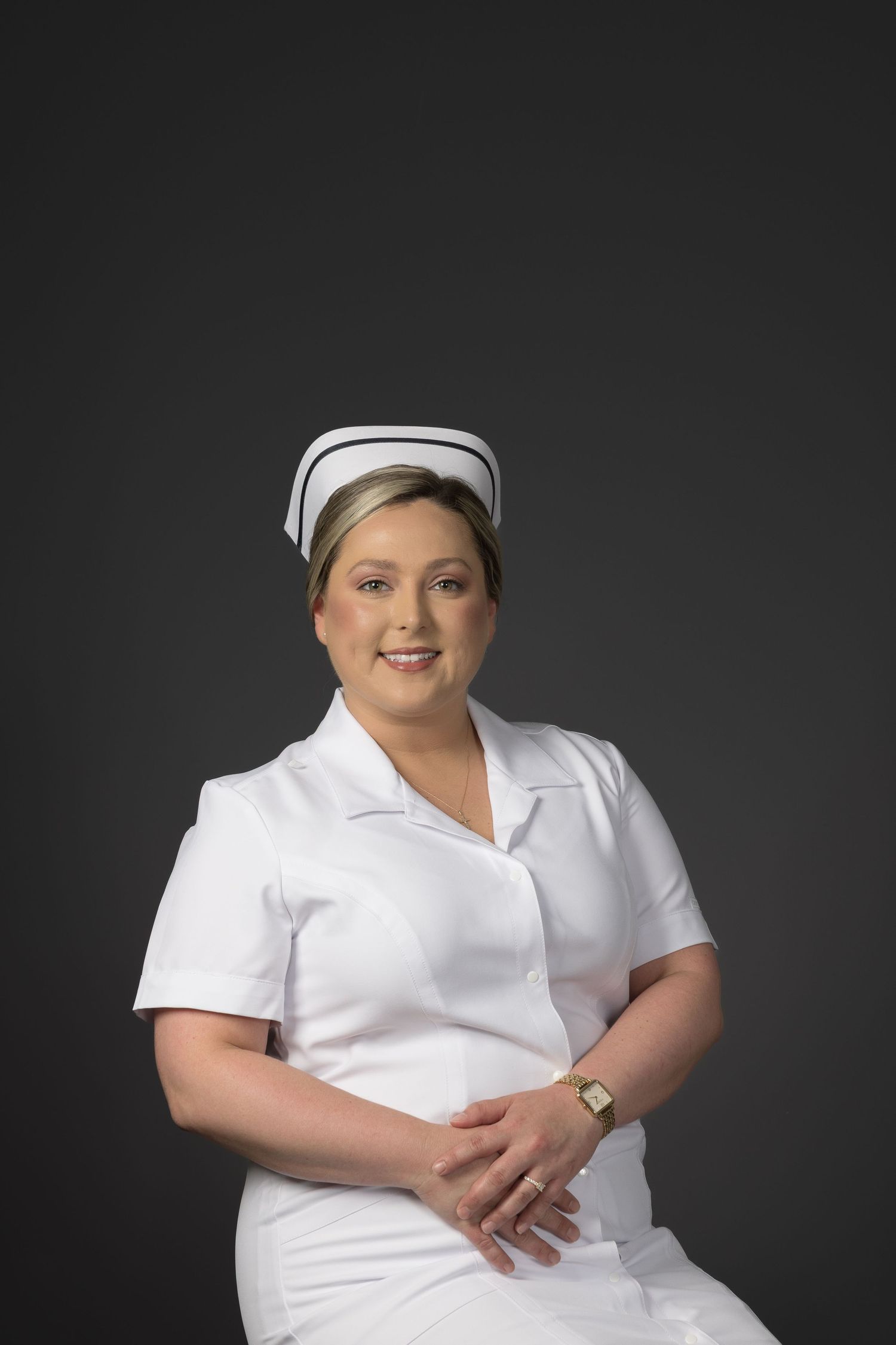 Female nurse wearing white uniform and cap sitting with hands folded against gray background.