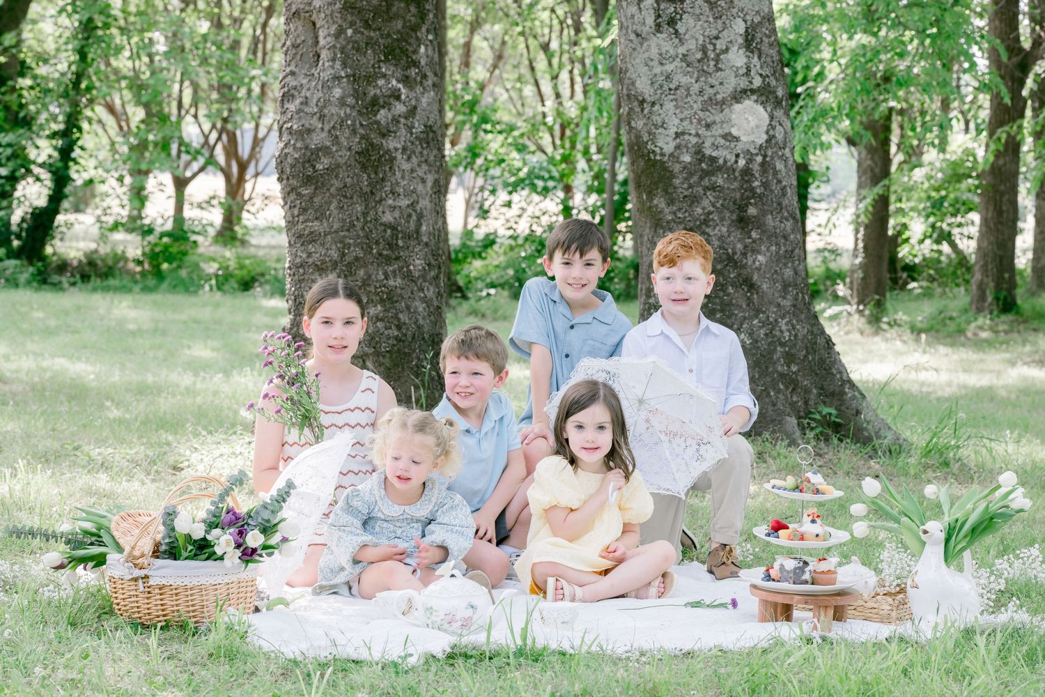 Six children pose for a spring picnic photo on a white blanket under large trees in a lush green park.