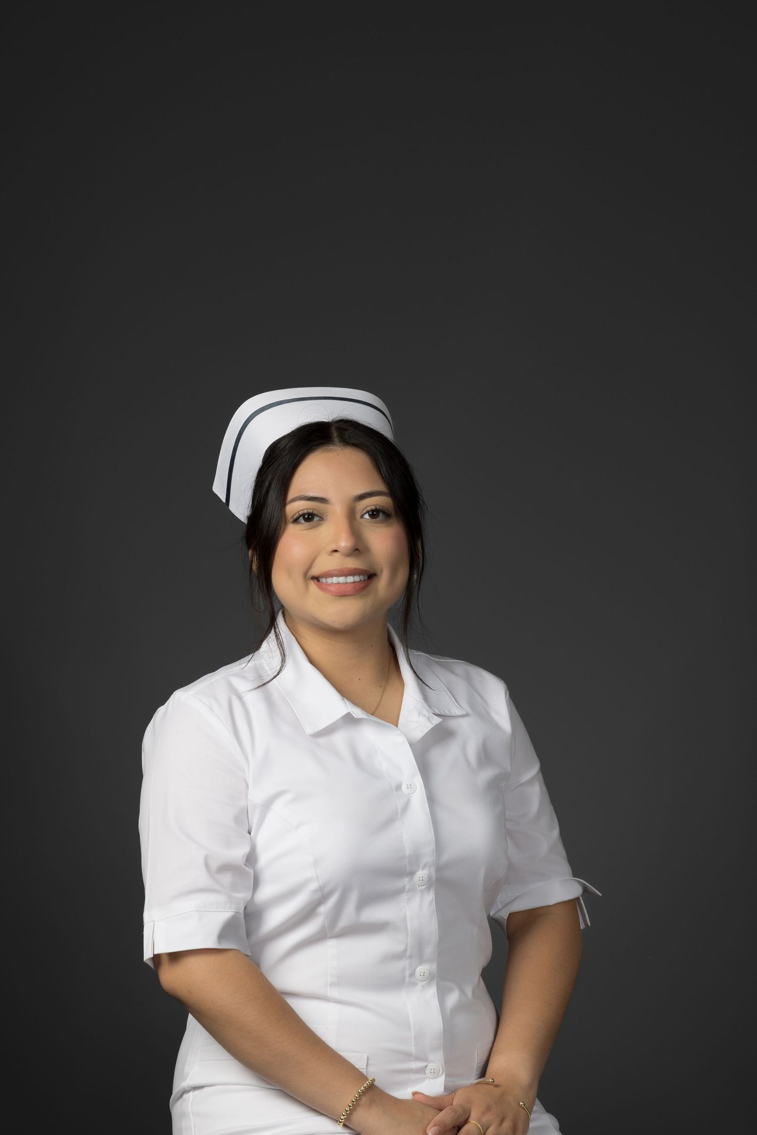 Smiling female nurse in white uniform and cap posing against a dark gray background.