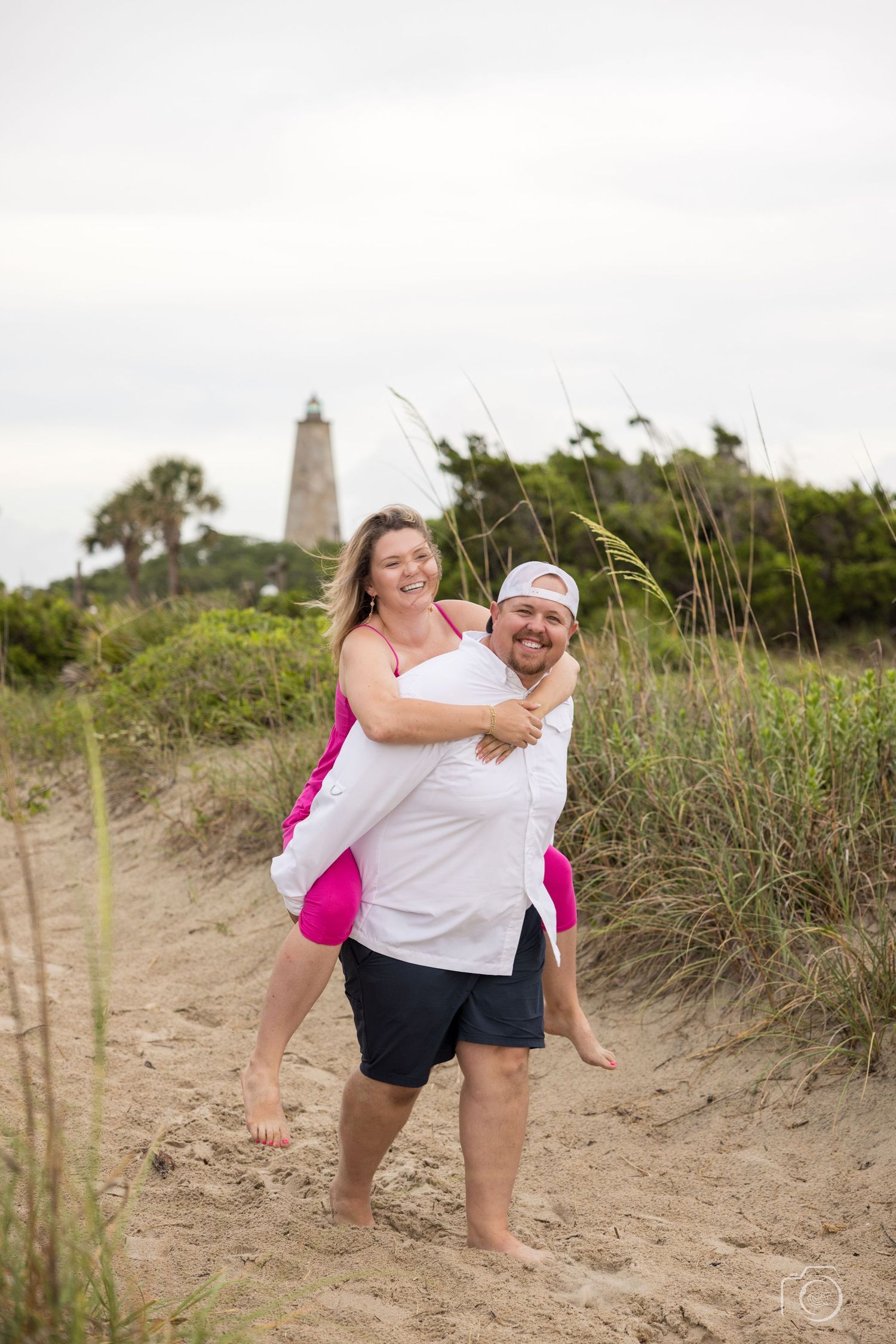 Playful beach photoshoot on sandy dunes with tall grass and lighthouse in background.