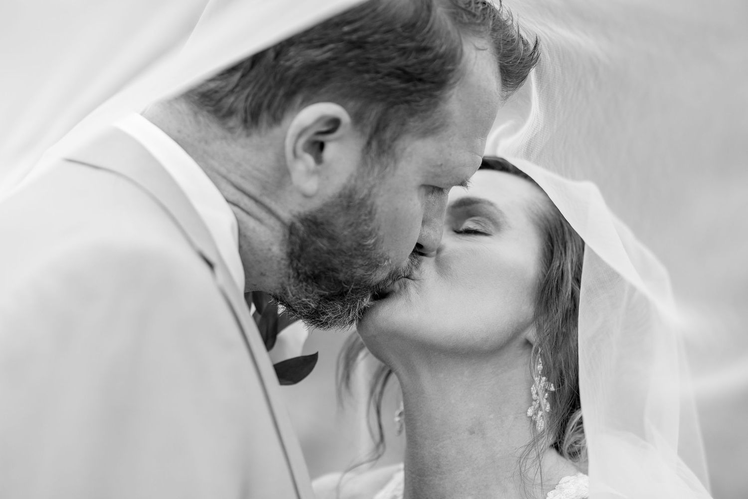 Black and white photo of a bride and groom sharing a romantic kiss under a wedding veil.