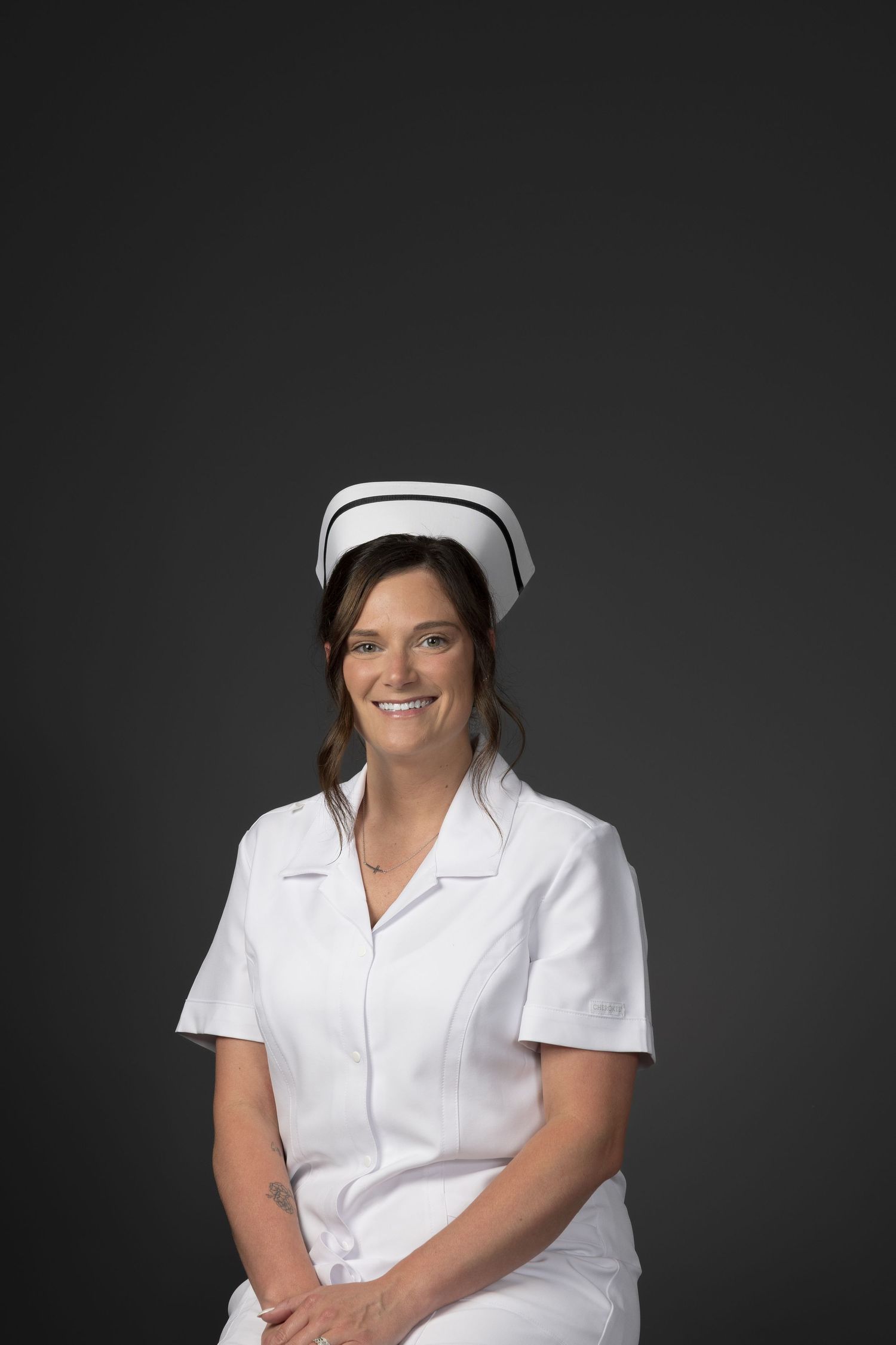 Smiling female nurse in white uniform and cap poses against dark gray background.