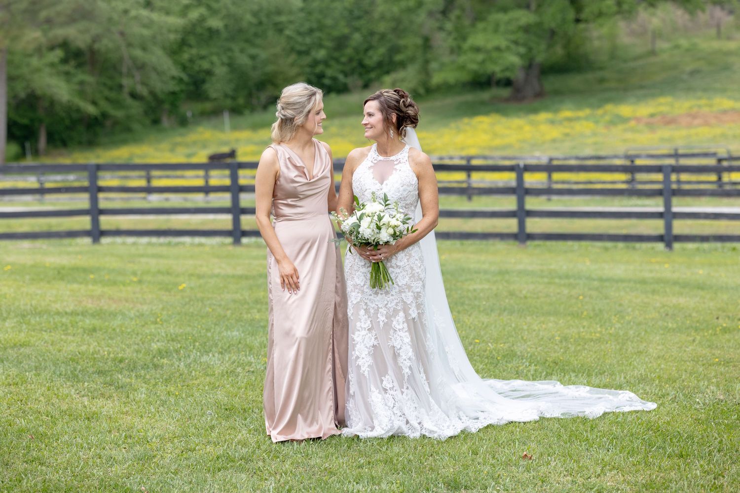 Bride in lace gown holding bouquet stands with bridesmaid in blush dress on green lawn near wooden fence.