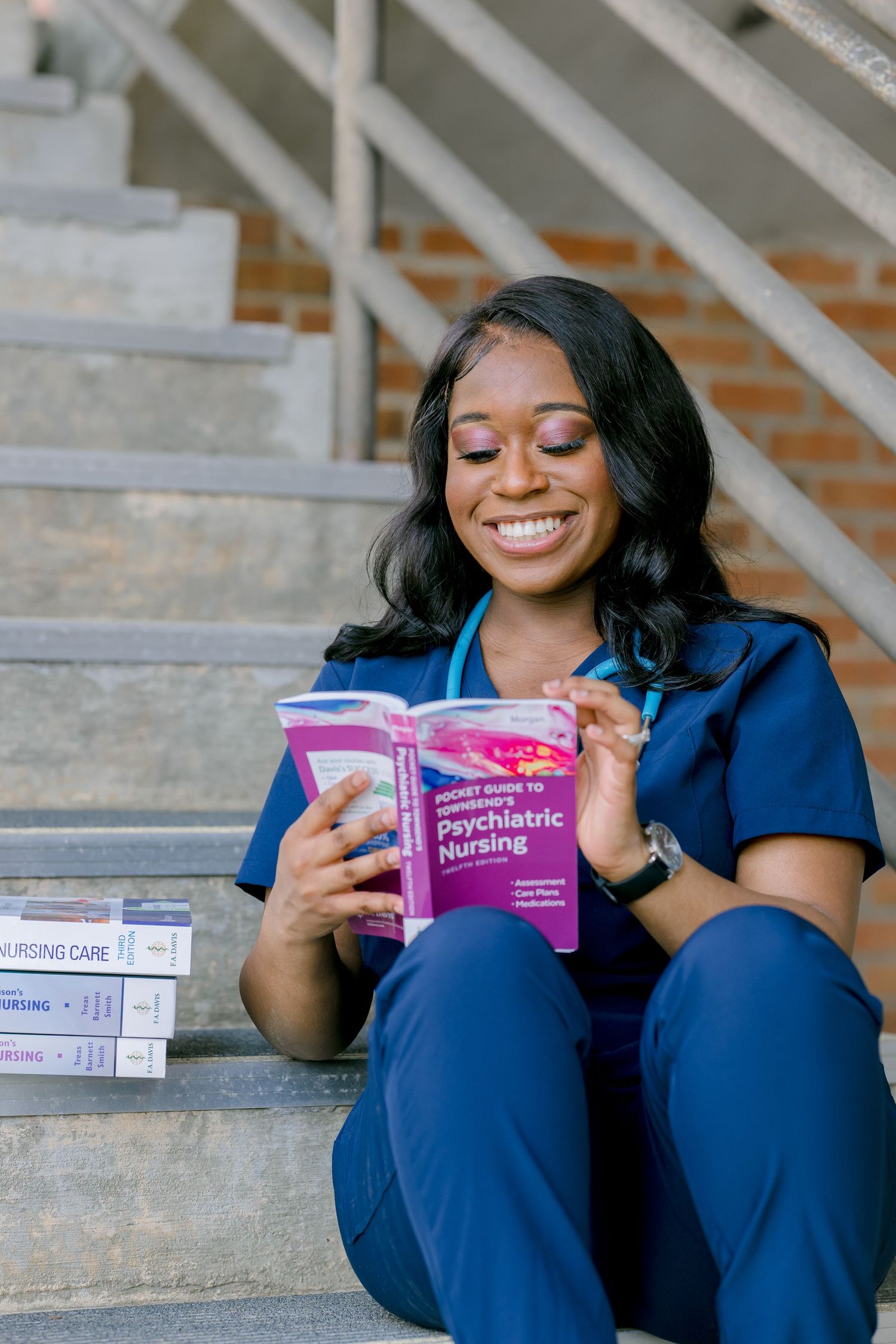 A student in blue scrubs sits on concrete steps studying from nursing textbooks and materials.