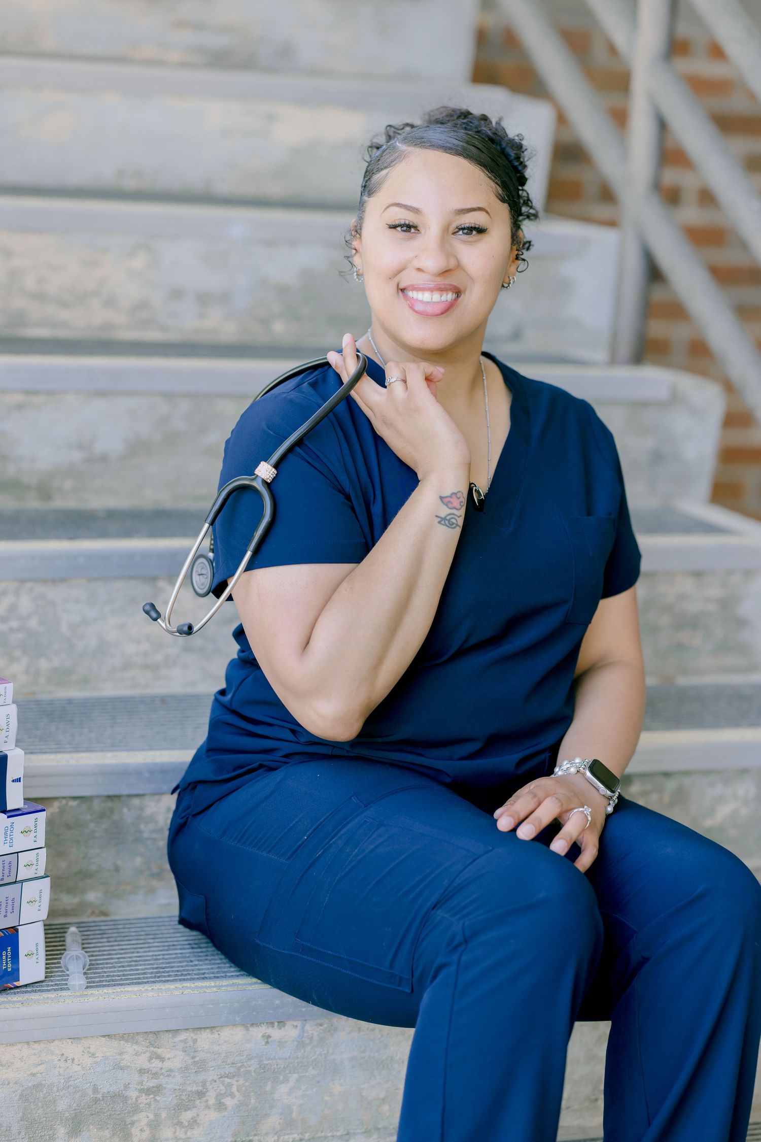 Smiling nurse in navy blue scrubs sits on steps holding a stethoscope, with medical supplies beside her.