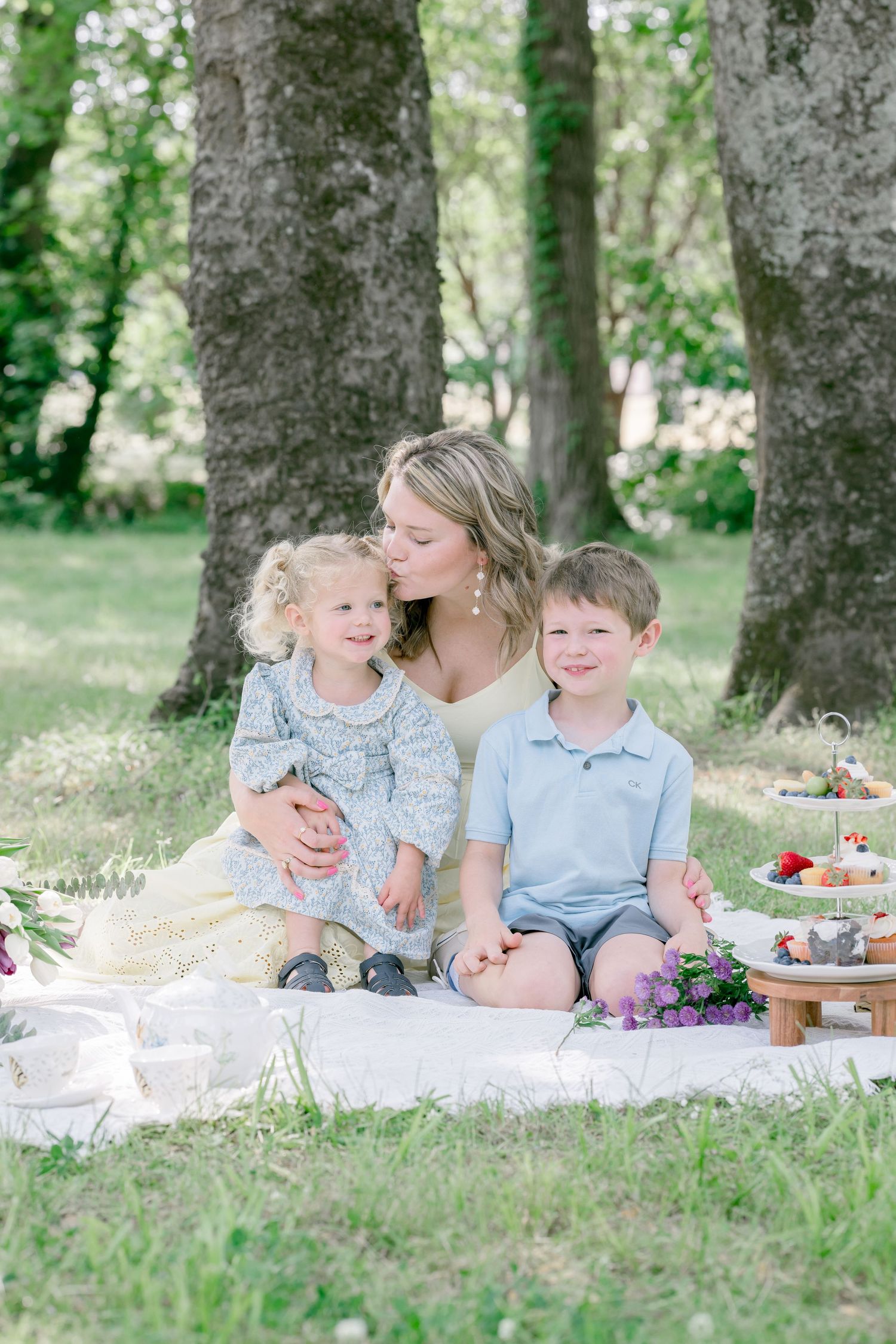 A mother kisses her toddler daughter while her young son smiles during an outdoor picnic photo session.
