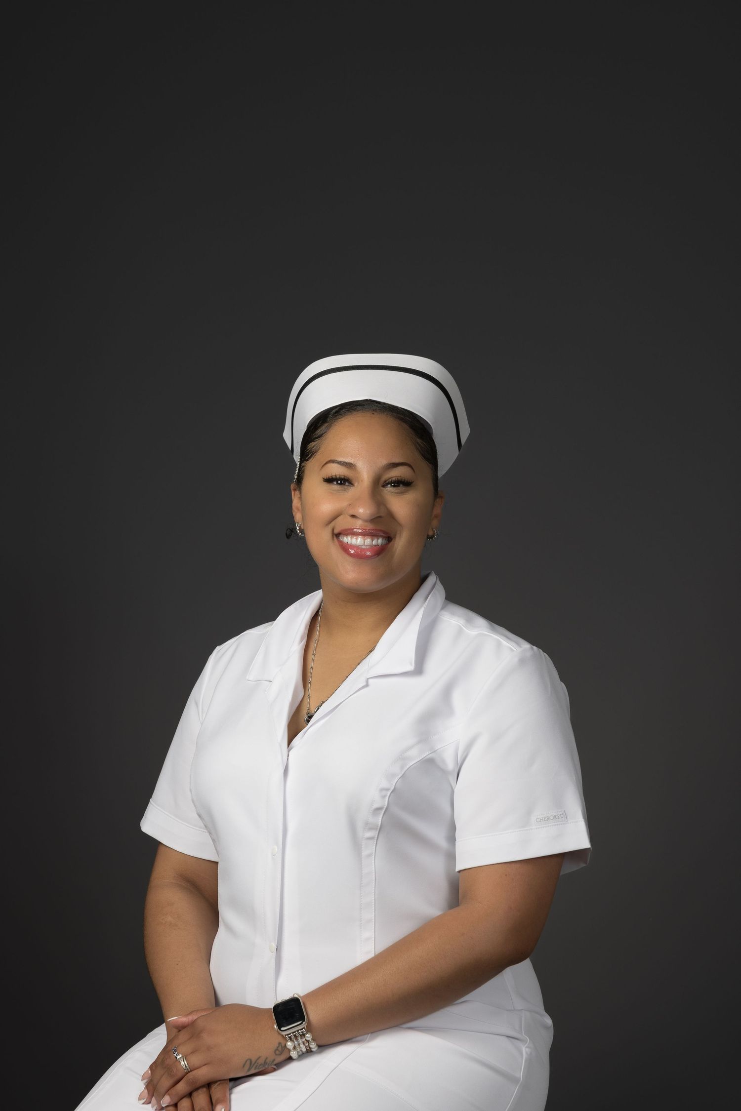 Smiling female nurse in white uniform and traditional cap posed against gray background.