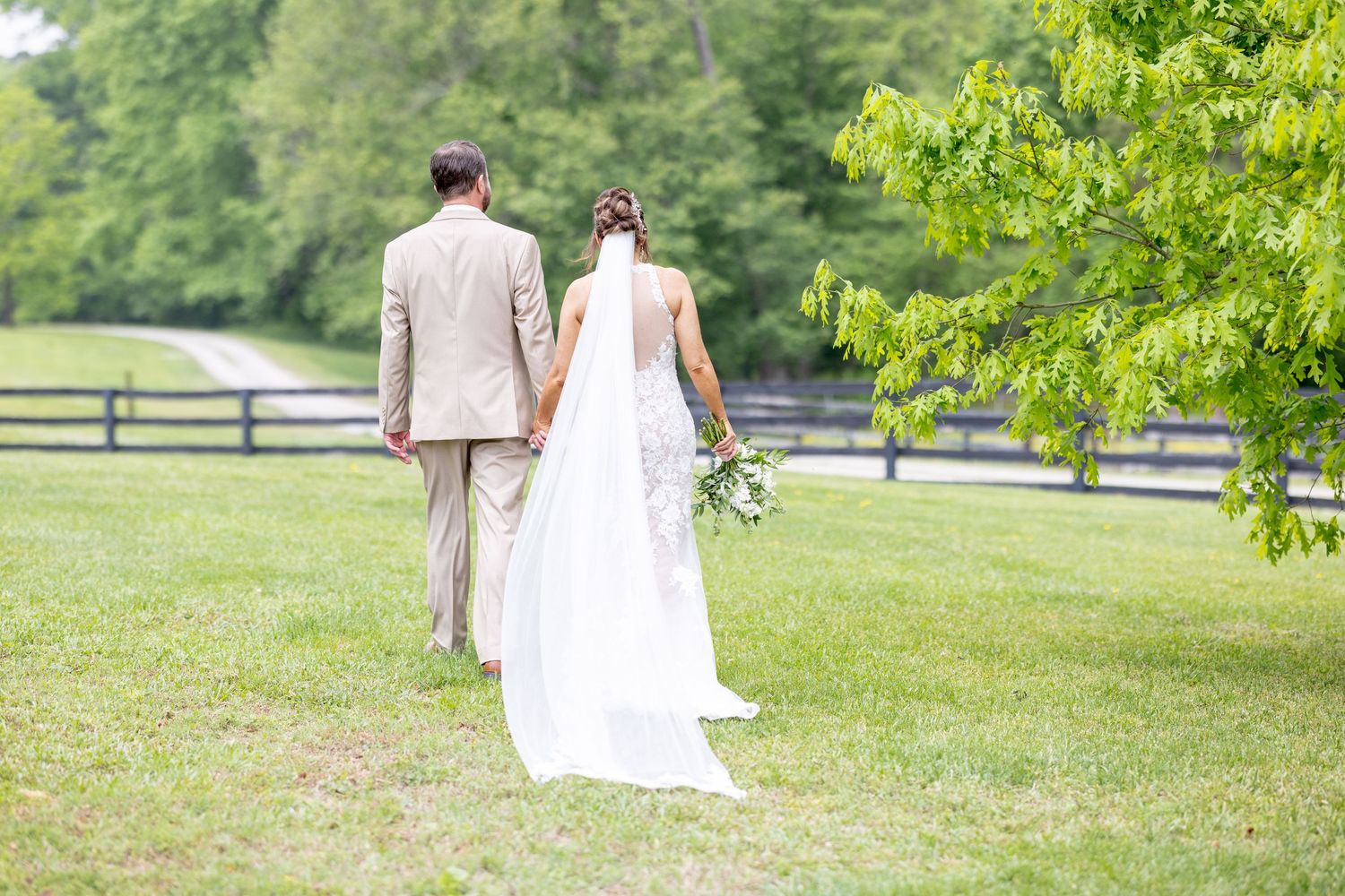 Bride and groom walking hand in hand across lush green lawn at outdoor wedding venue with wooden fence.