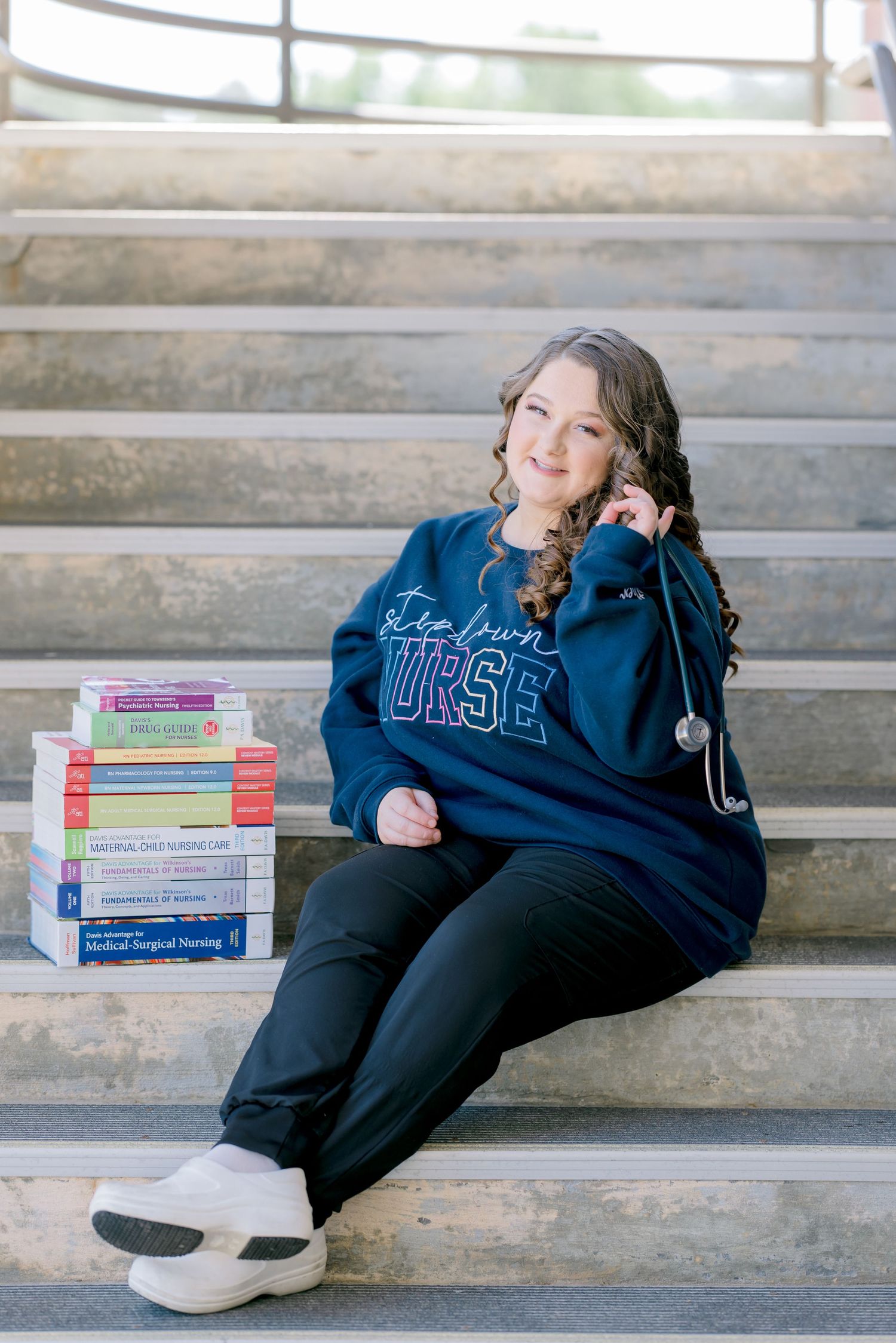 A nursing student sits on stone steps beside stacked medical textbooks, wearing a navy nurse sweatshirt and stethoscope.